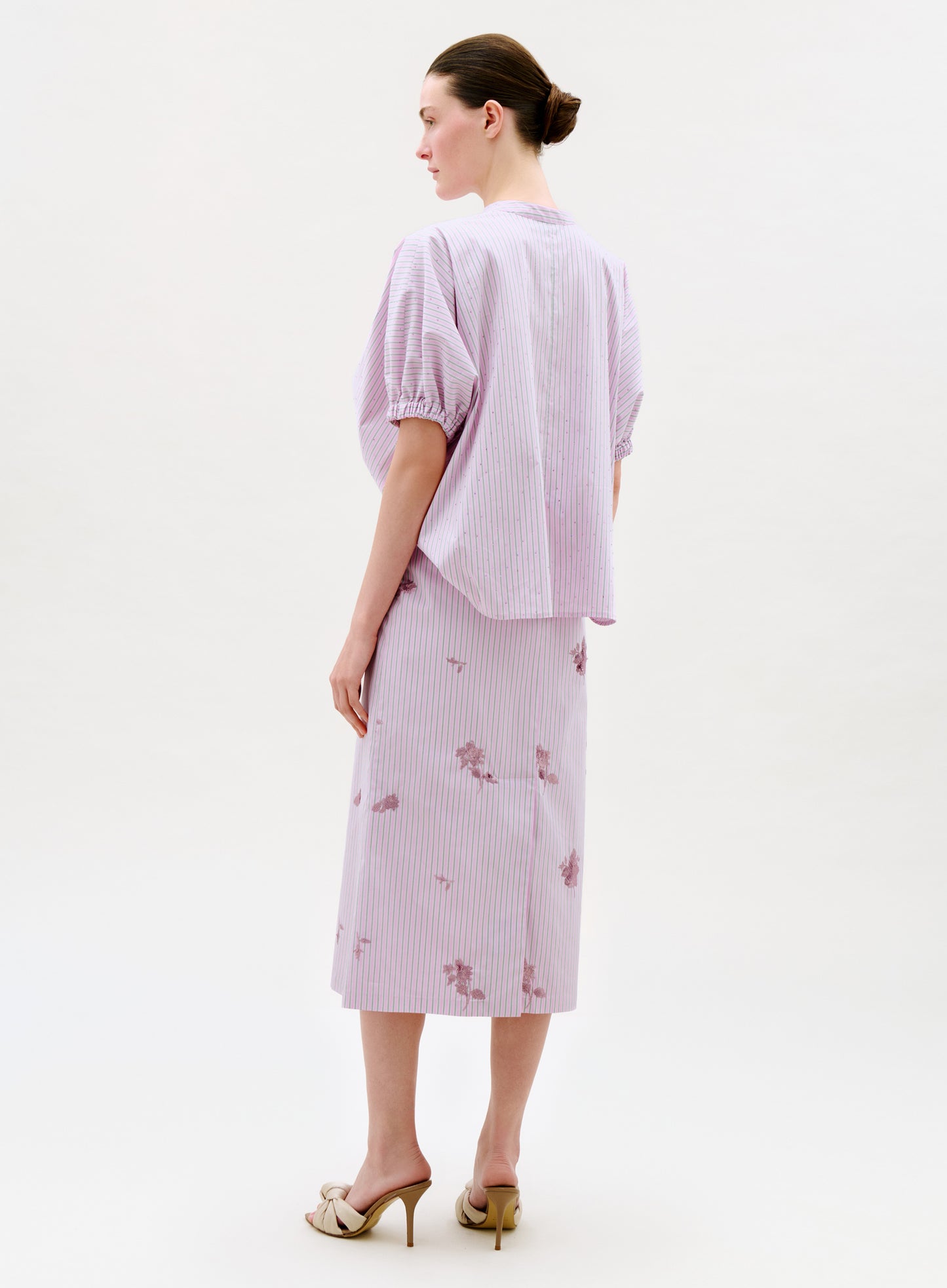 A woman faces away, wearing a light purple short-sleeve blouse paired with the Atira Skirt— a matching midi skirt featuring subtle floral patterns. She has her hair tied back and cream high-heeled sandals, set against a plain background.