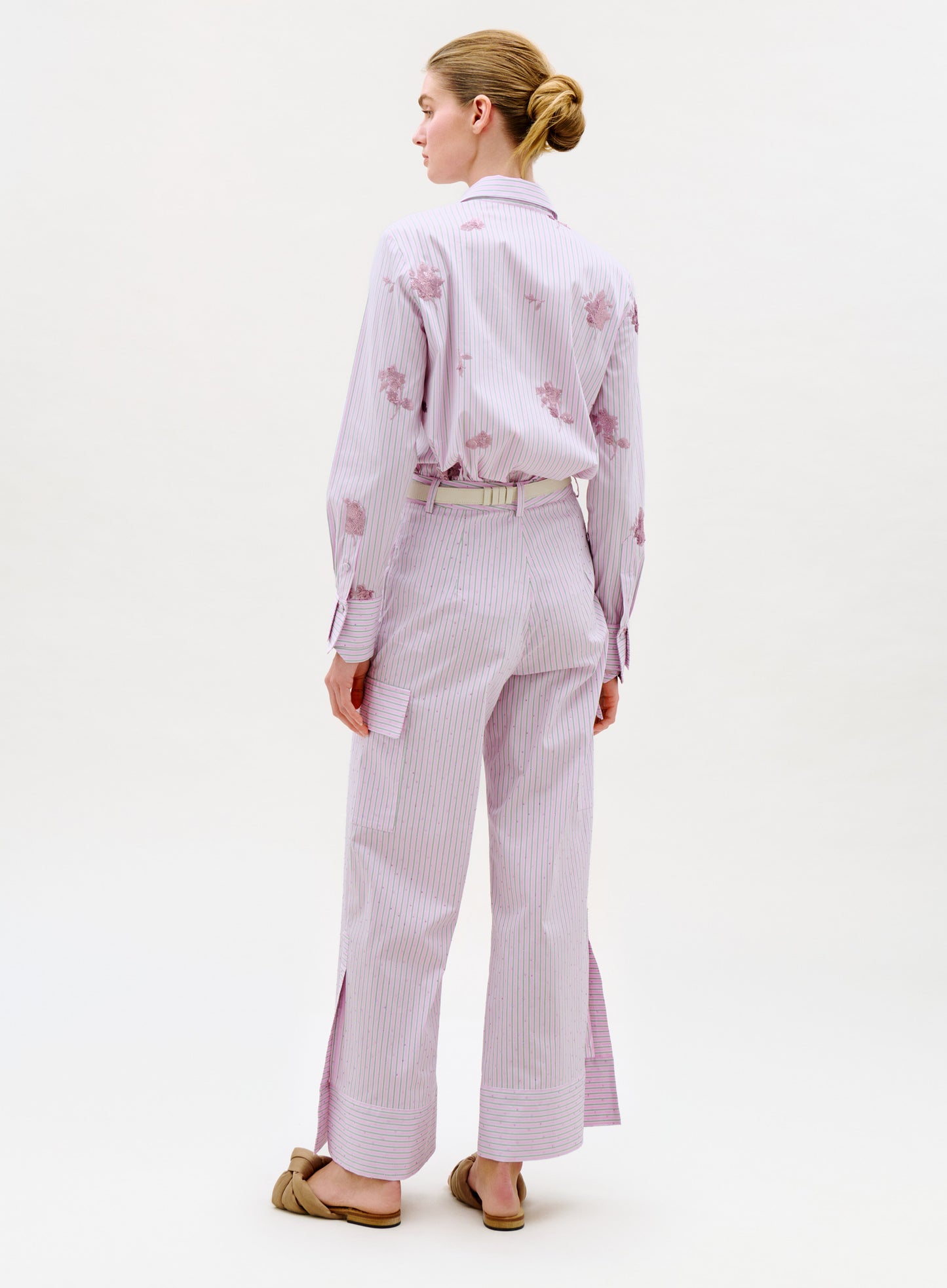 A woman with her hair in a bun wears the Falak Pant—pale pink and white stripes with floral patterns, wide legs, and slits at the hem—along with a matching top and tan slip-on shoes. She stands facing away.
