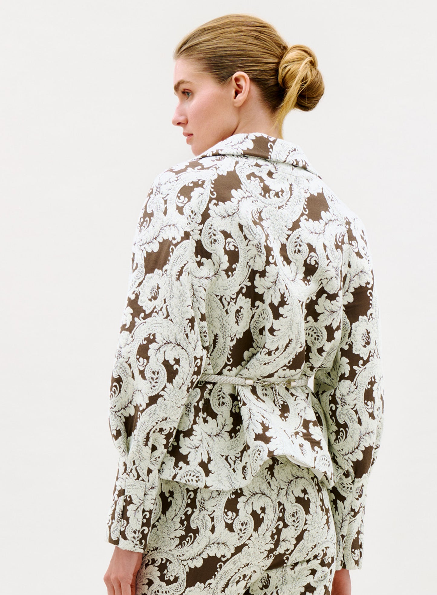 A woman with blonde hair in a low bun is seen from behind and slightly to the side, wearing the Noureen Jacket—a brown and white paisley-patterned suit jacket with a belt—set against a plain light background.
