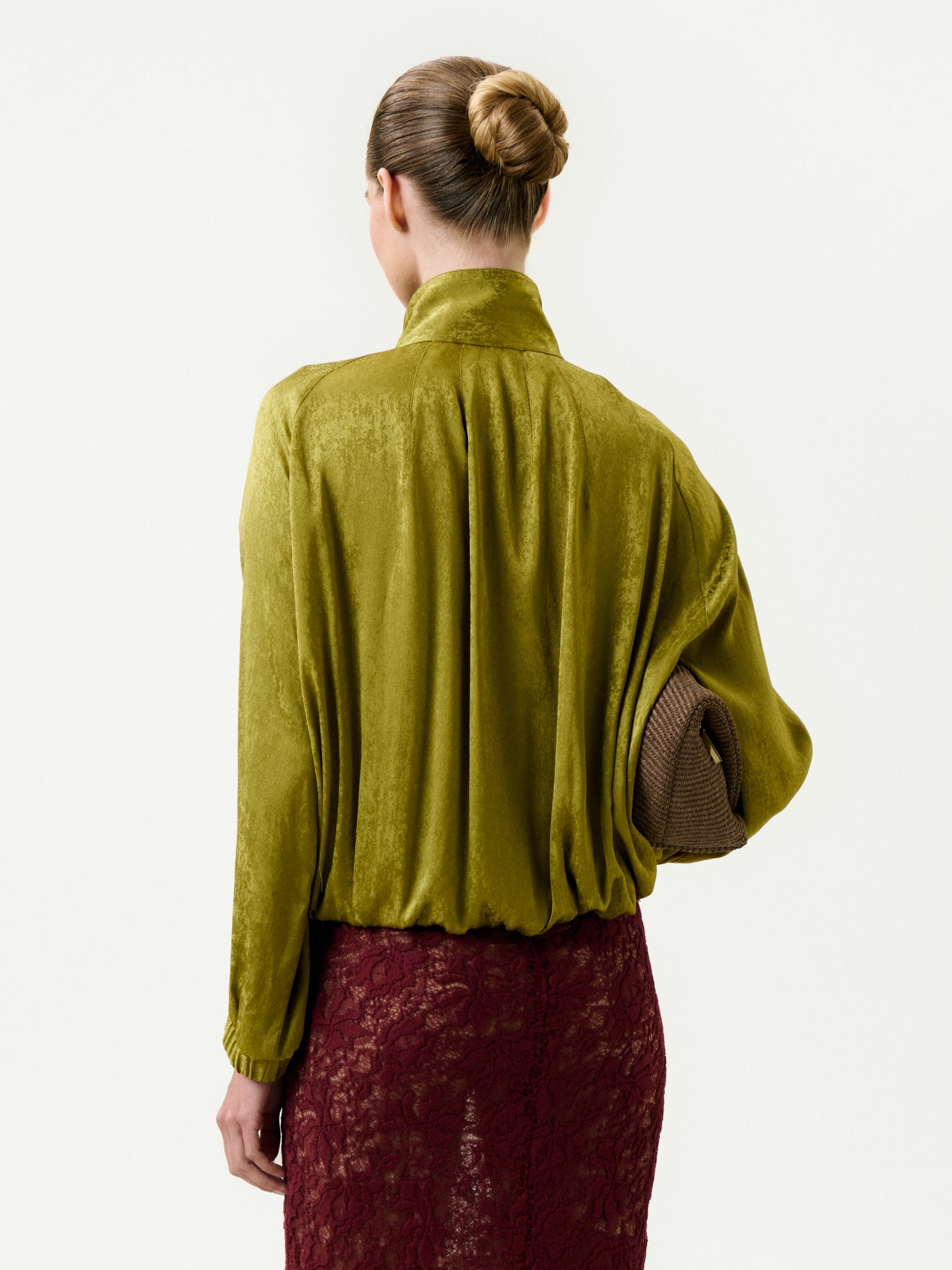 A person with hair in a neat bun is seen from behind, wearing the Jhene Jacket and a burgundy lace skirt, holding a textured brown clutch under one arm against a plain light background.