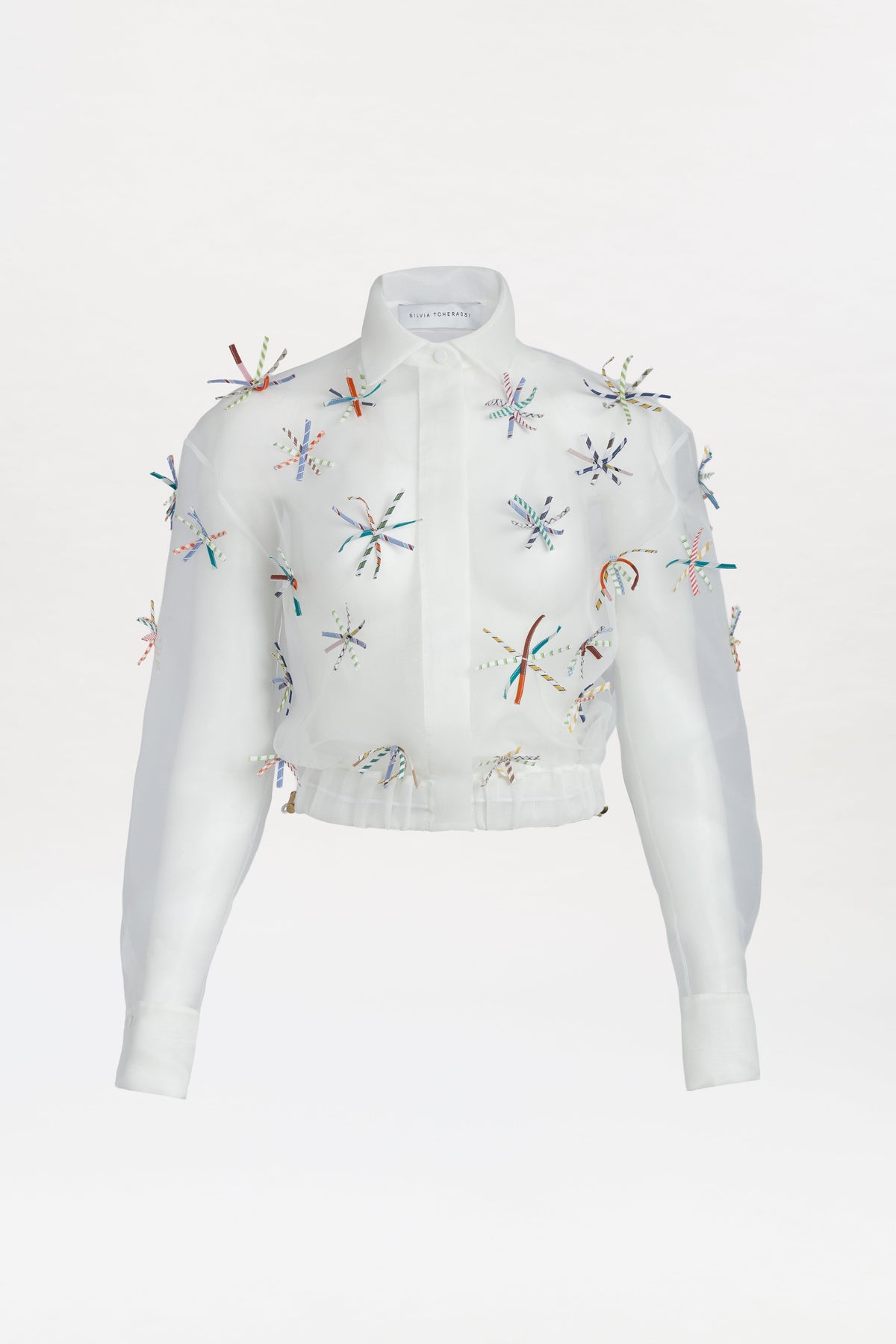 The Katniss Jacket is a white, long-sleeve collared piece featuring colorful embroidered starburst patterns and small beads scattered across the slightly sheer fabric. It has a button-down front for easy wear.