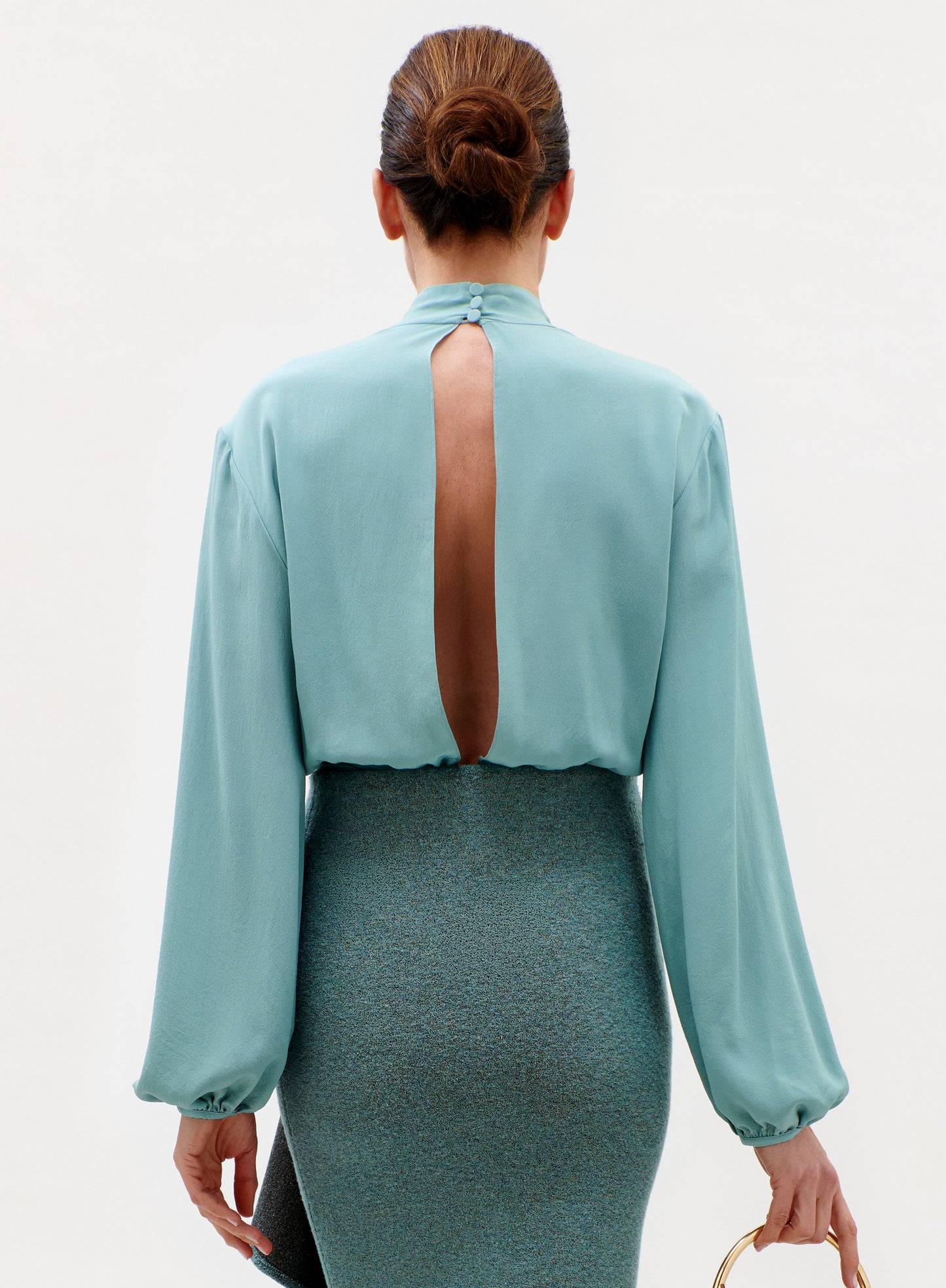 A woman stands facing away, wearing the Delfina Blouse—a light blue, long-sleeved top with an open back slit—paired with a textured high-waisted skirt and her hair in a neat bun.