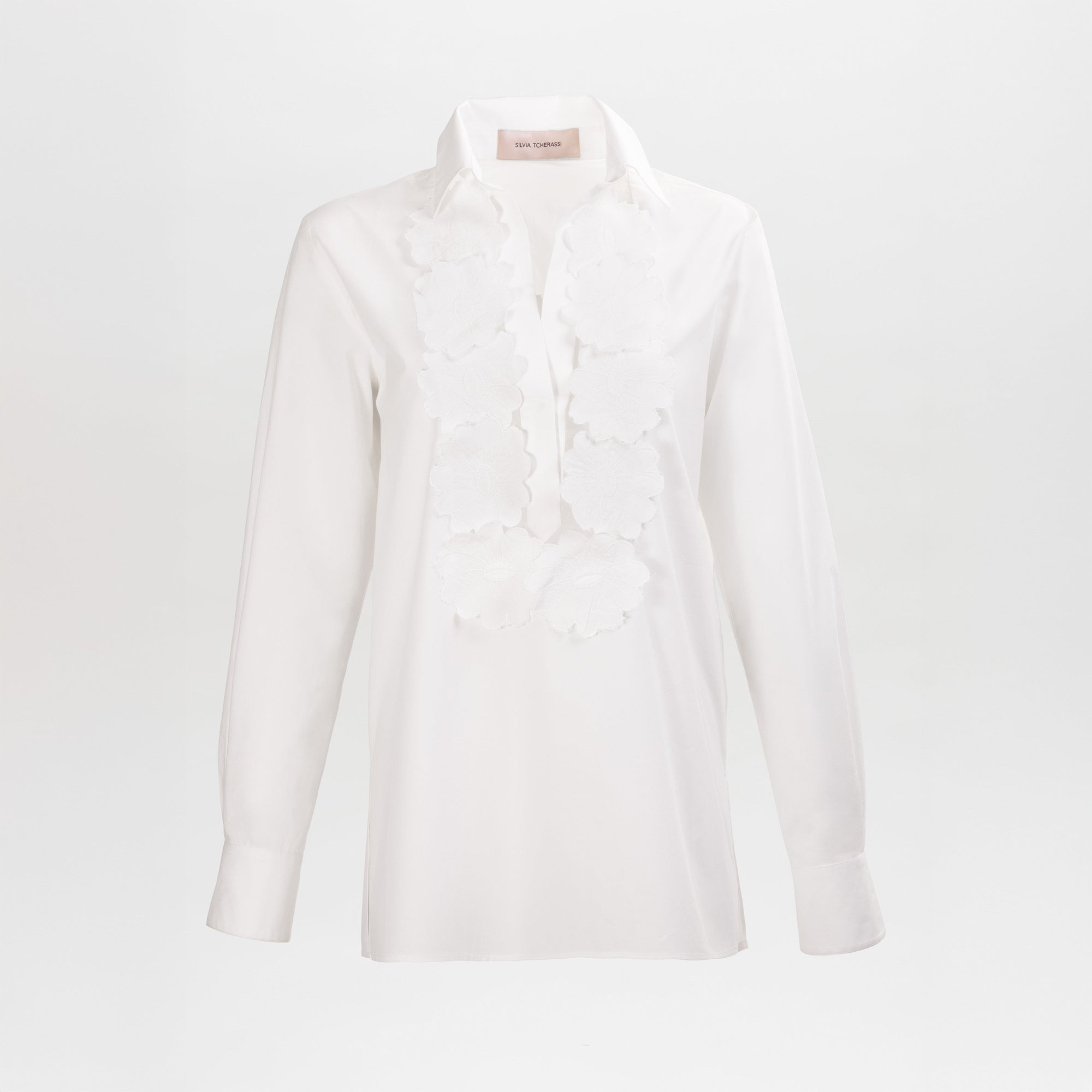 The Karole Blouse is a long-sleeve white blouse with a collar and large floral appliqué details along the front placket, shown against a plain white background.