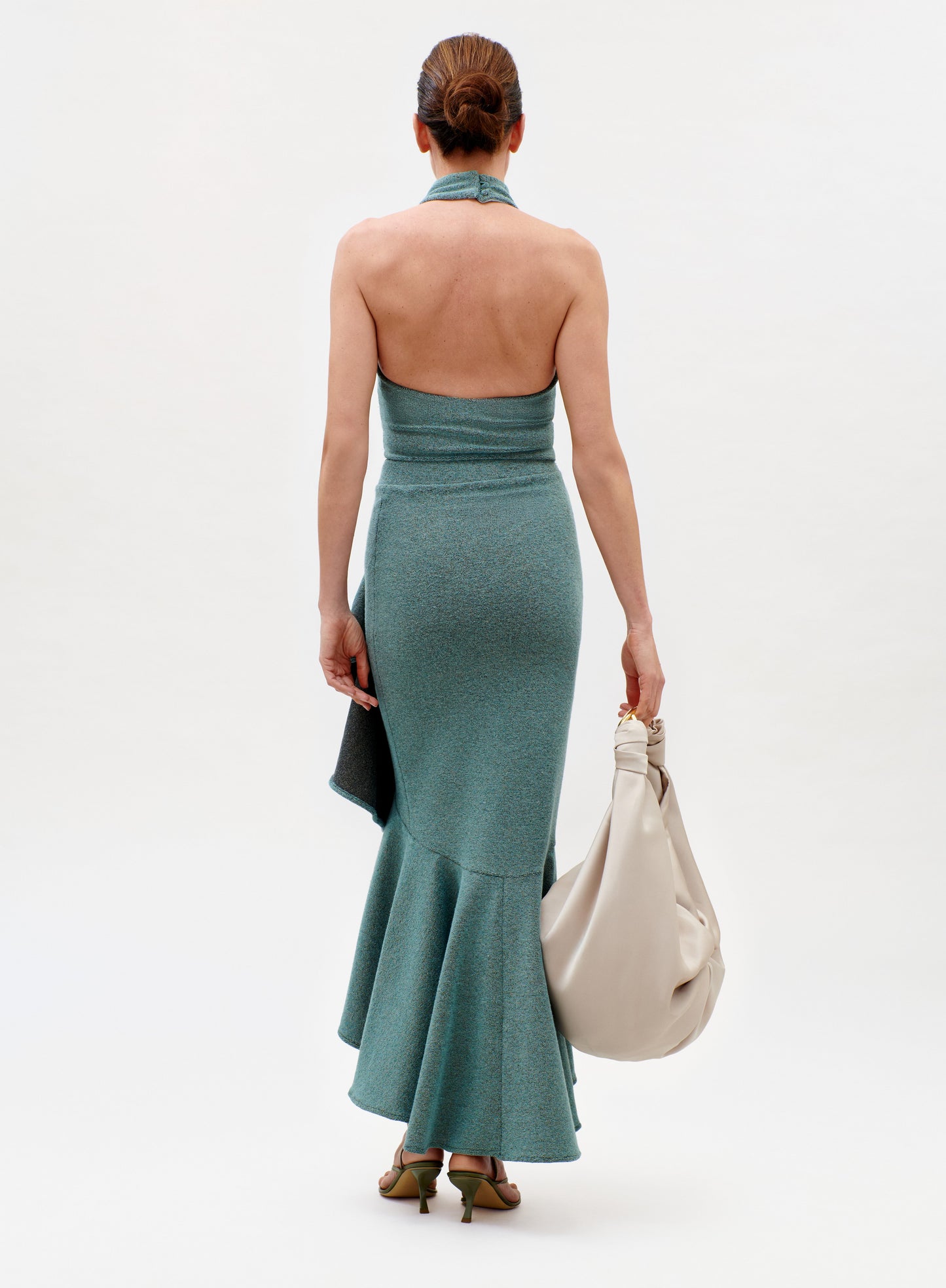 A woman faces away wearing the Bambolera Skirt, a teal, fitted halter-neck dress with a flared hem. She carries a large off-white knotted handbag, wears green heels, and has brown hair styled in a neat bun against a white background.
