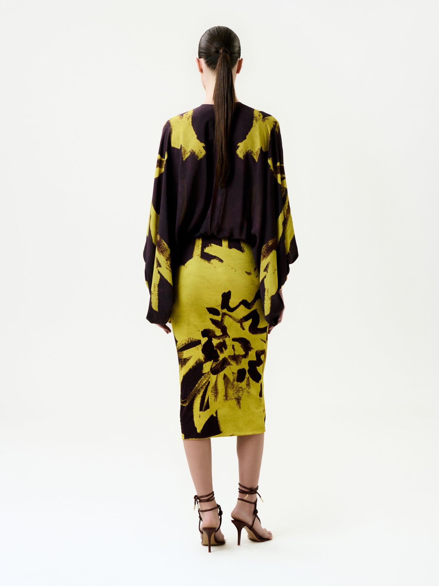 A woman with long dark hair in a ponytail is seen from behind wearing the Kumiko Dress, a knee-length piece with batwing sleeves, crafted from luxurious Italian fabric featuring bold yellow and dark purple abstract florals.