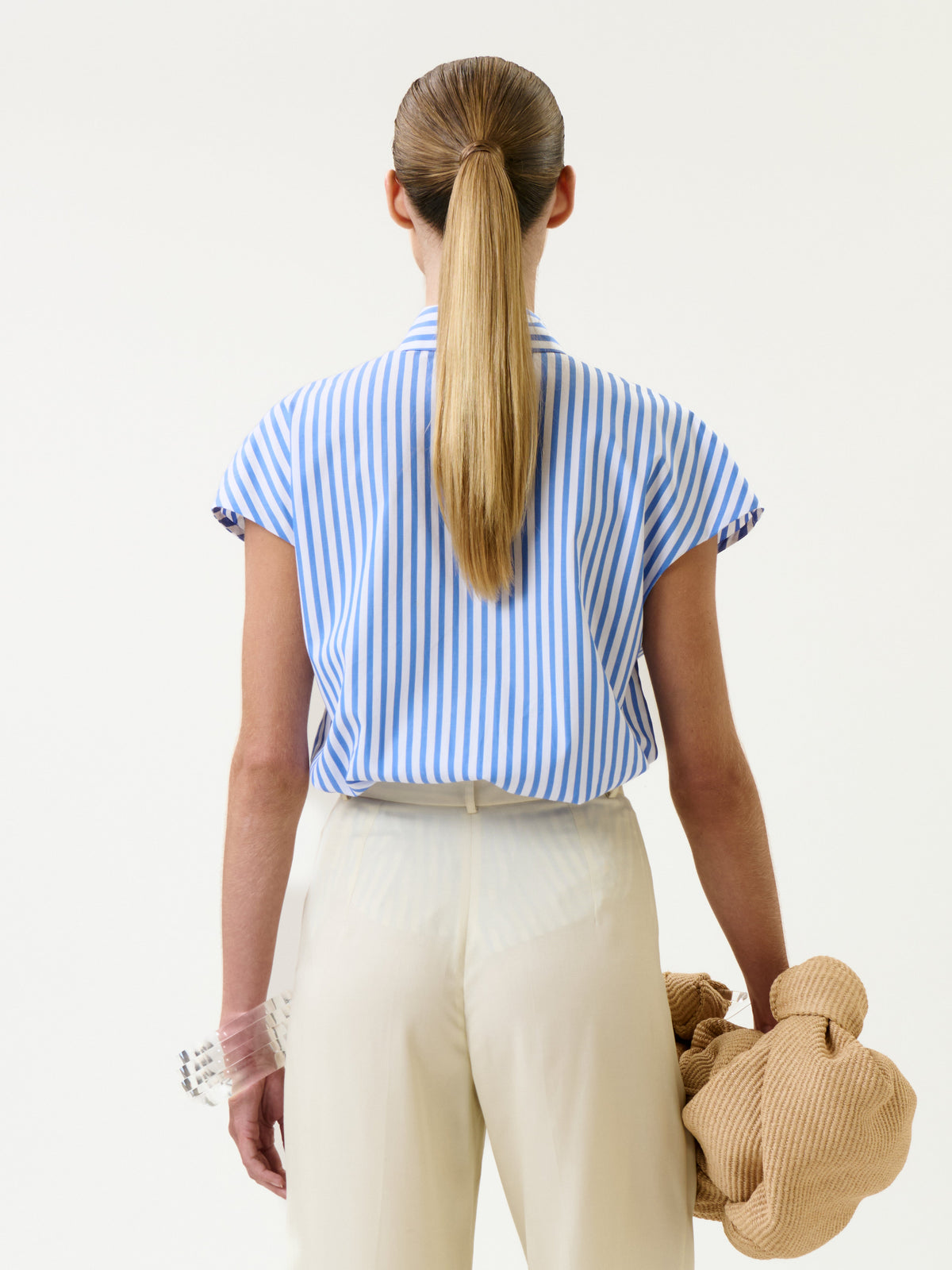 A woman with a sleek ponytail is wearing the Maribel Shirt and cream pants. She is facing away and holding a textured beige object in her left hand, against a plain light background.