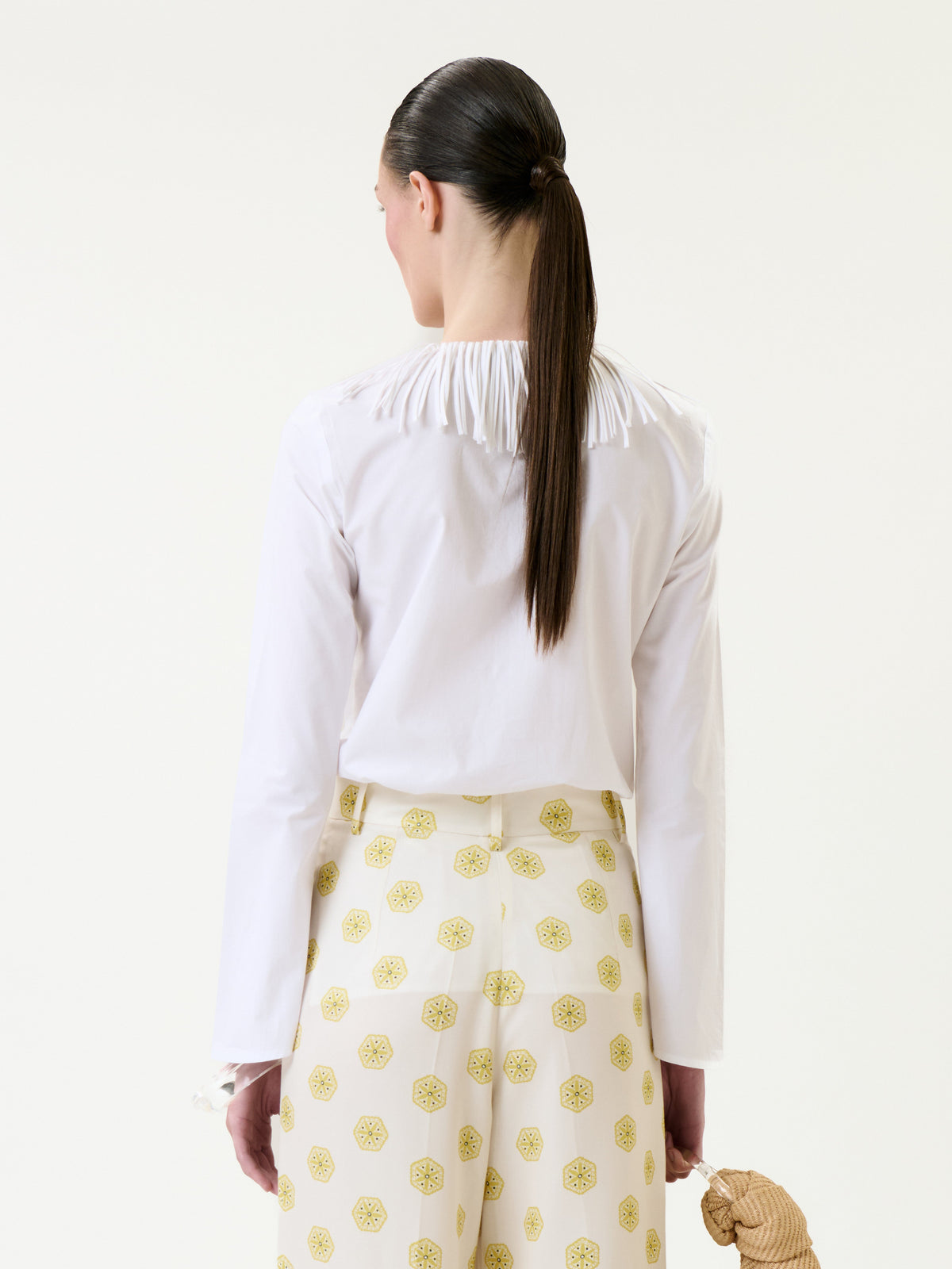 A person with long dark hair in a ponytail is shown from the back against a plain light background, wearing the white Zena Shirt and yellow patterned pants while holding a straw hat.