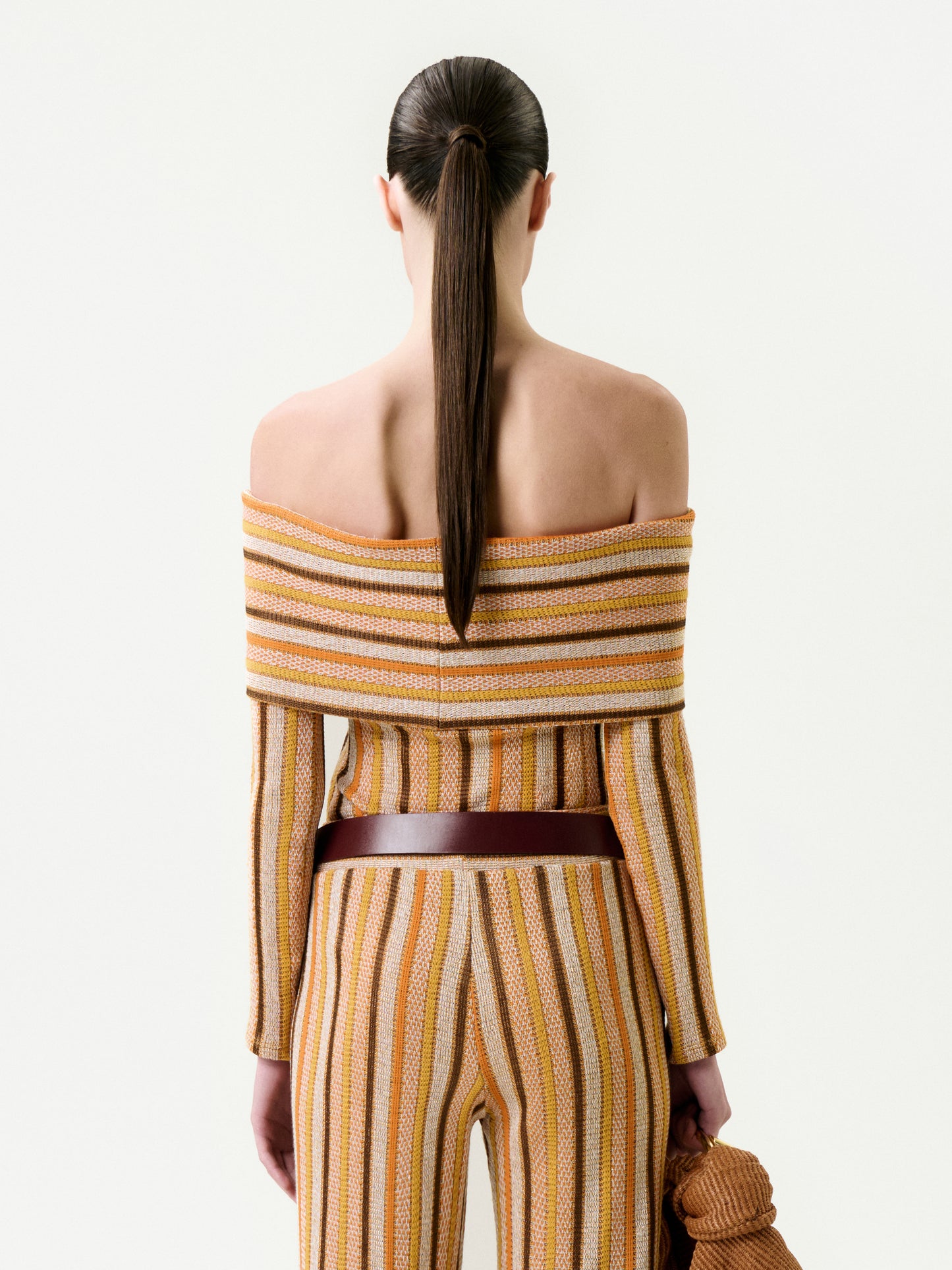 A woman with long brown hair in a ponytail faces away, wearing the Maitenea Top—an off-the-shoulder, ribbed-knit striped piece in brown, yellow, and white Italian fabric—and holds a matching accessory in her left hand.