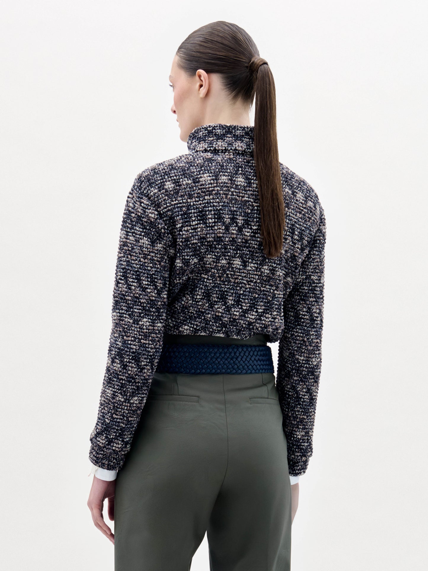 A person stands against a plain background wearing the Zalta Jacket, olive green pants, and a wide blue belt with a large gold buckle.