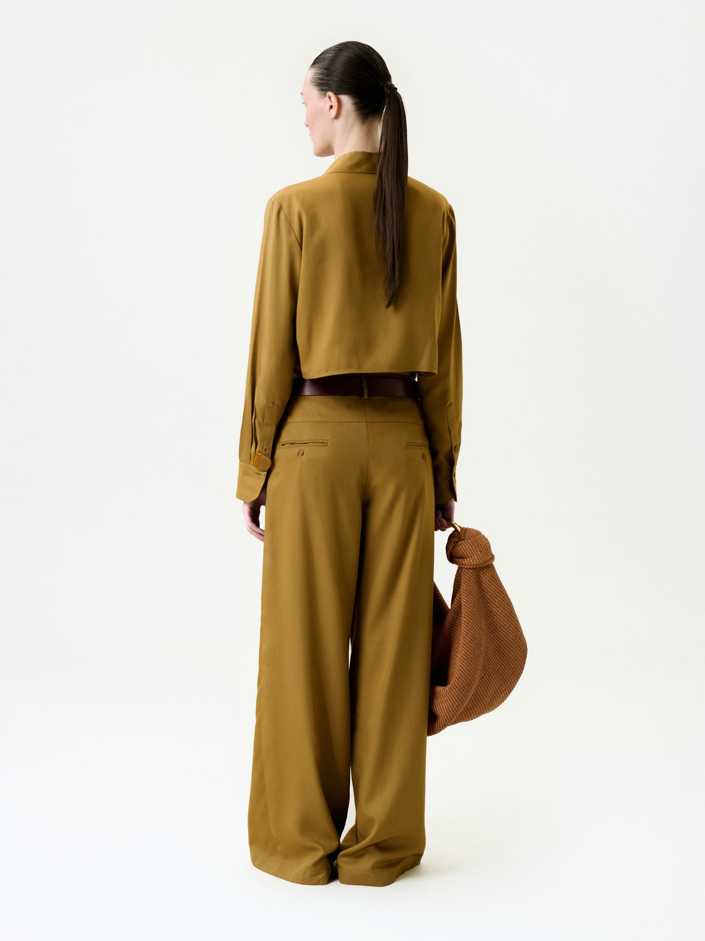 A woman with dark hair tied back stands against a white background, wearing relaxed fit wide-leg Opal Pants and a mustard yellow long-sleeve top. She holds a textured brown bag in her left hand, facing away from the camera.