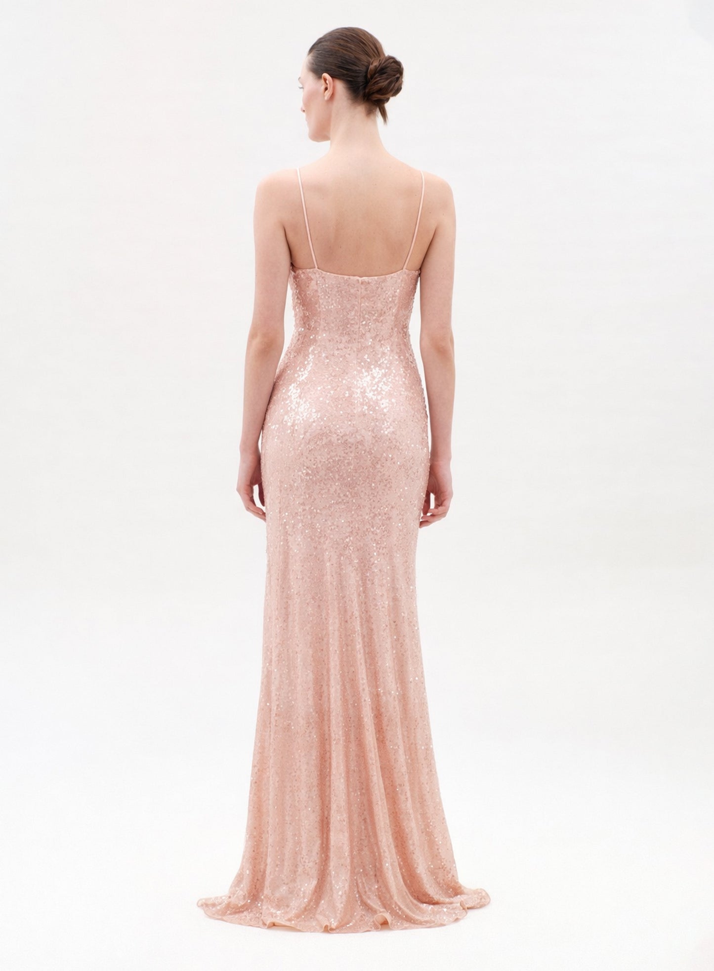 A woman stands facing away, wearing the Ibona Dress—a long, blush pink sequin gown with thin spaghetti straps and a low back. Her hair is in a neat bun as the dress softly pools on the light-colored floor.