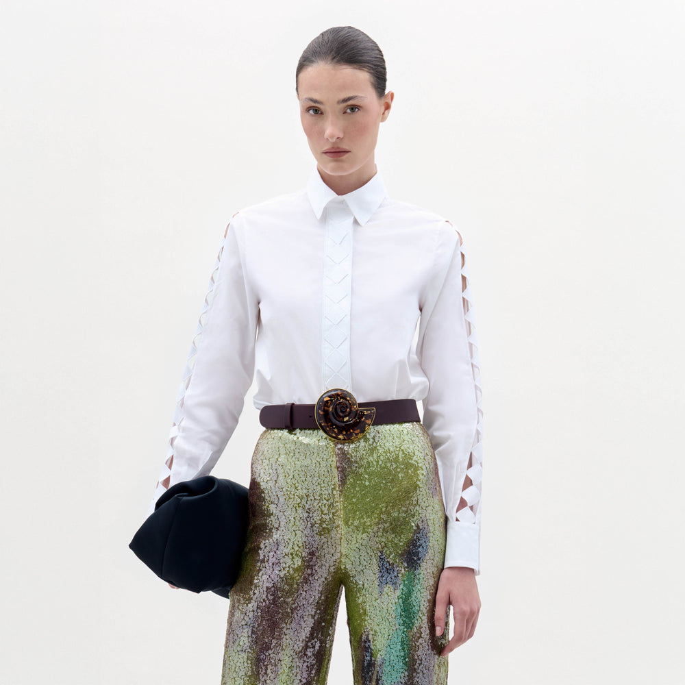 A woman stands against a plain background wearing the Idris Blouse with cut-out sleeves, paired with iridescent wide-leg pants, a large belt, and holding a folded black garment.