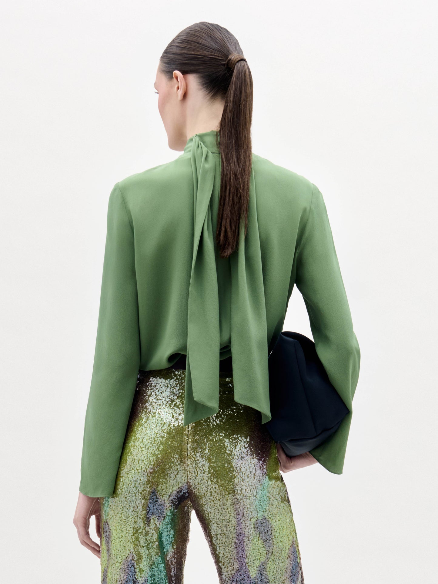 A woman poses against a plain background wearing the green Kelsey Blouse, sequined pants, a wide belt, and holding a black clutch.