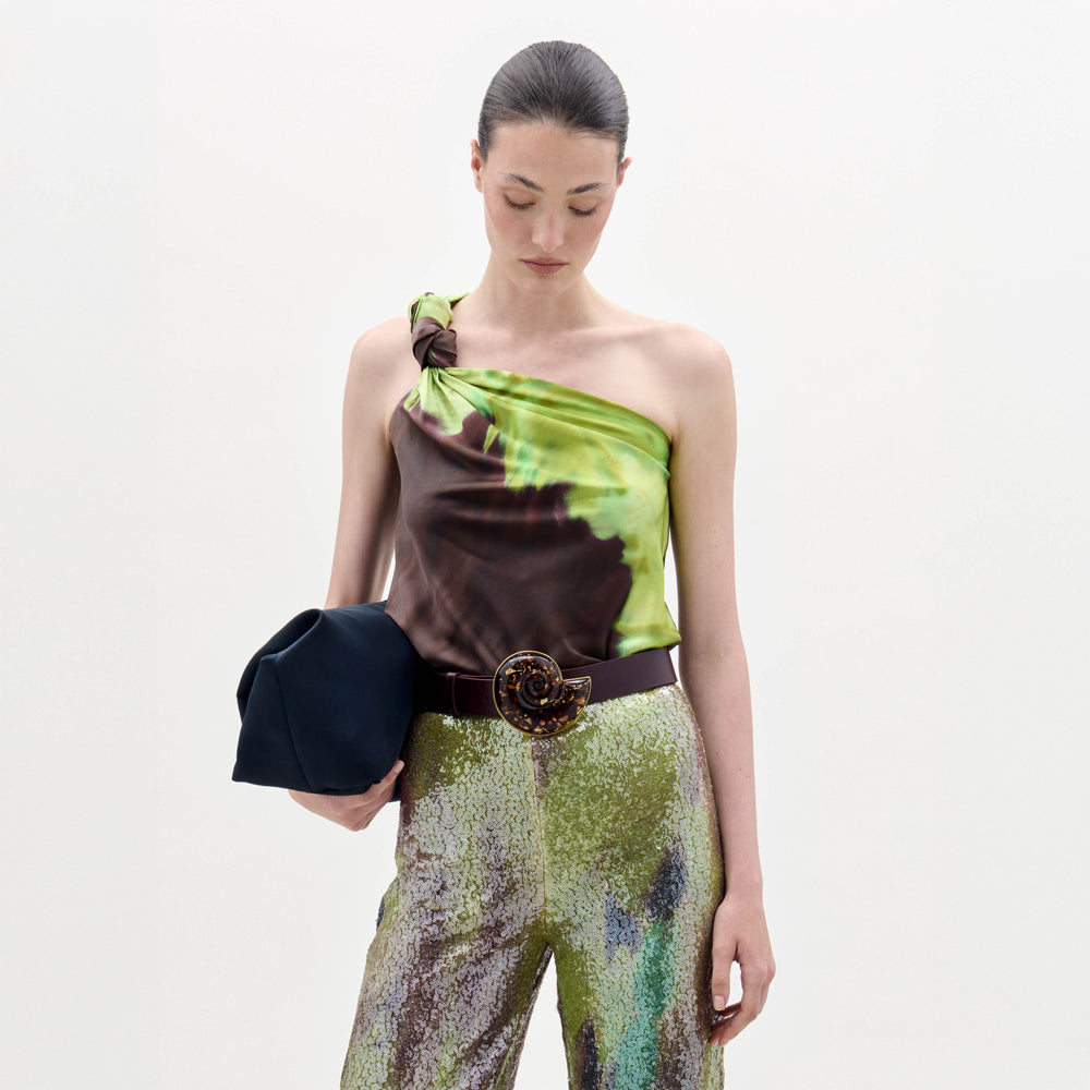 A woman stands against a plain background wearing the Yanet Blouse, a one-shoulder green and brown top, paired with sequined pants, a wide belt with a large buckle, and holding a black clutch.