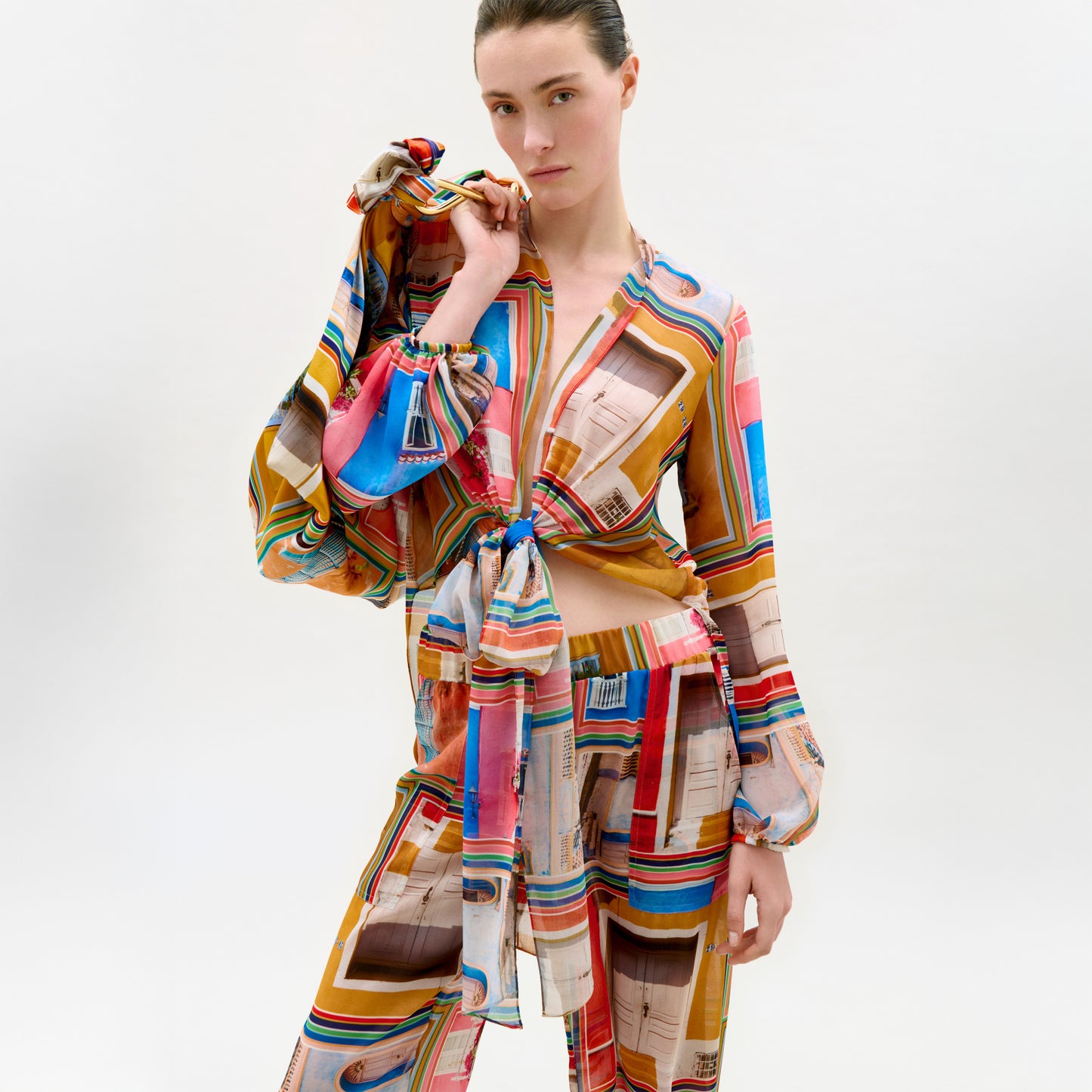 A woman poses against a white background wearing the Honey Blouse, an Italian fabric long-sleeve top with front tie detail, paired with matching loose, colorful patterned pants.