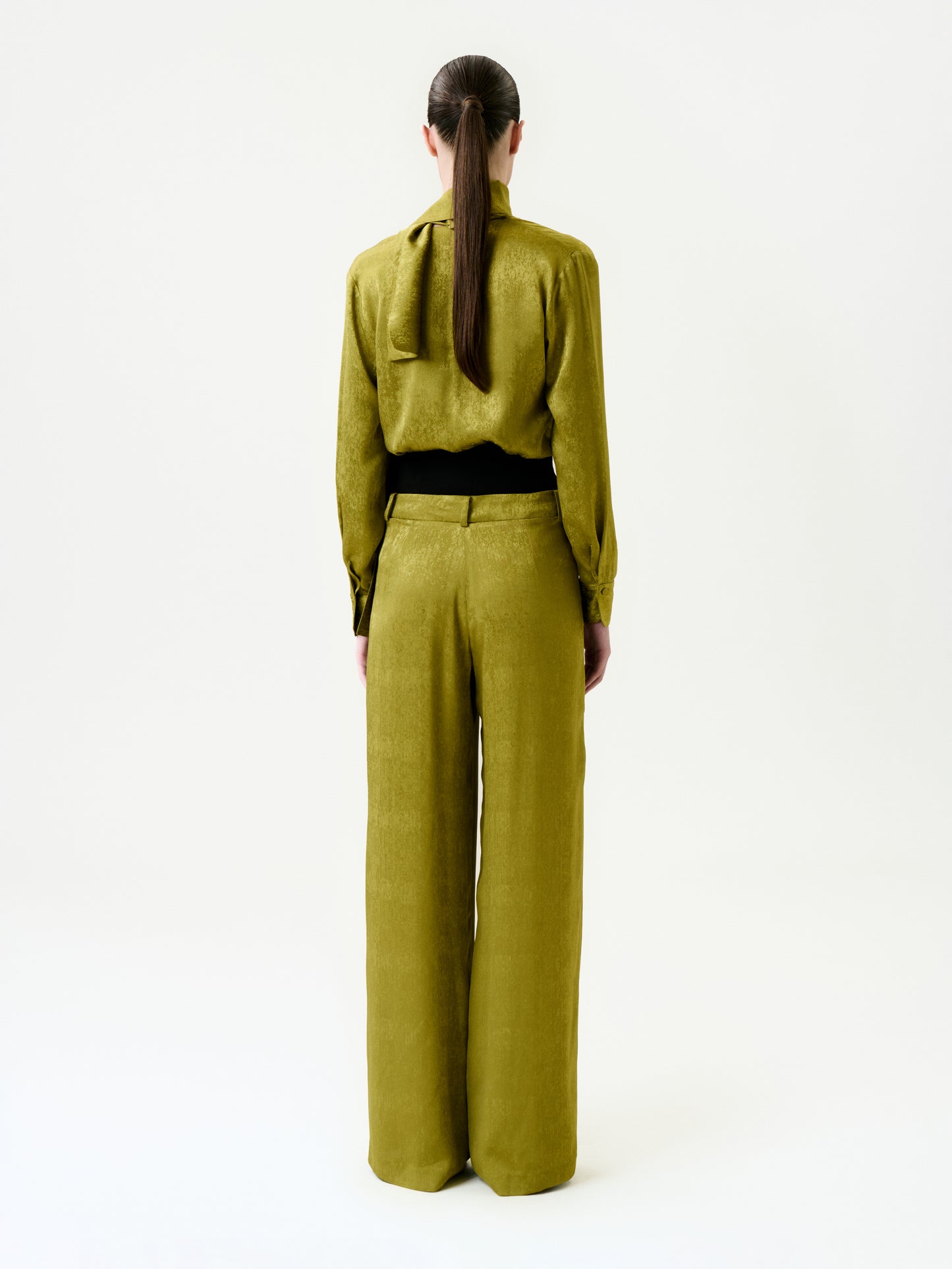 A person with long brown hair in a low ponytail stands facing away, wearing a shiny olive green blouse and the Karel Pant, wide-leg trousers made in Colombia, against a plain white background.