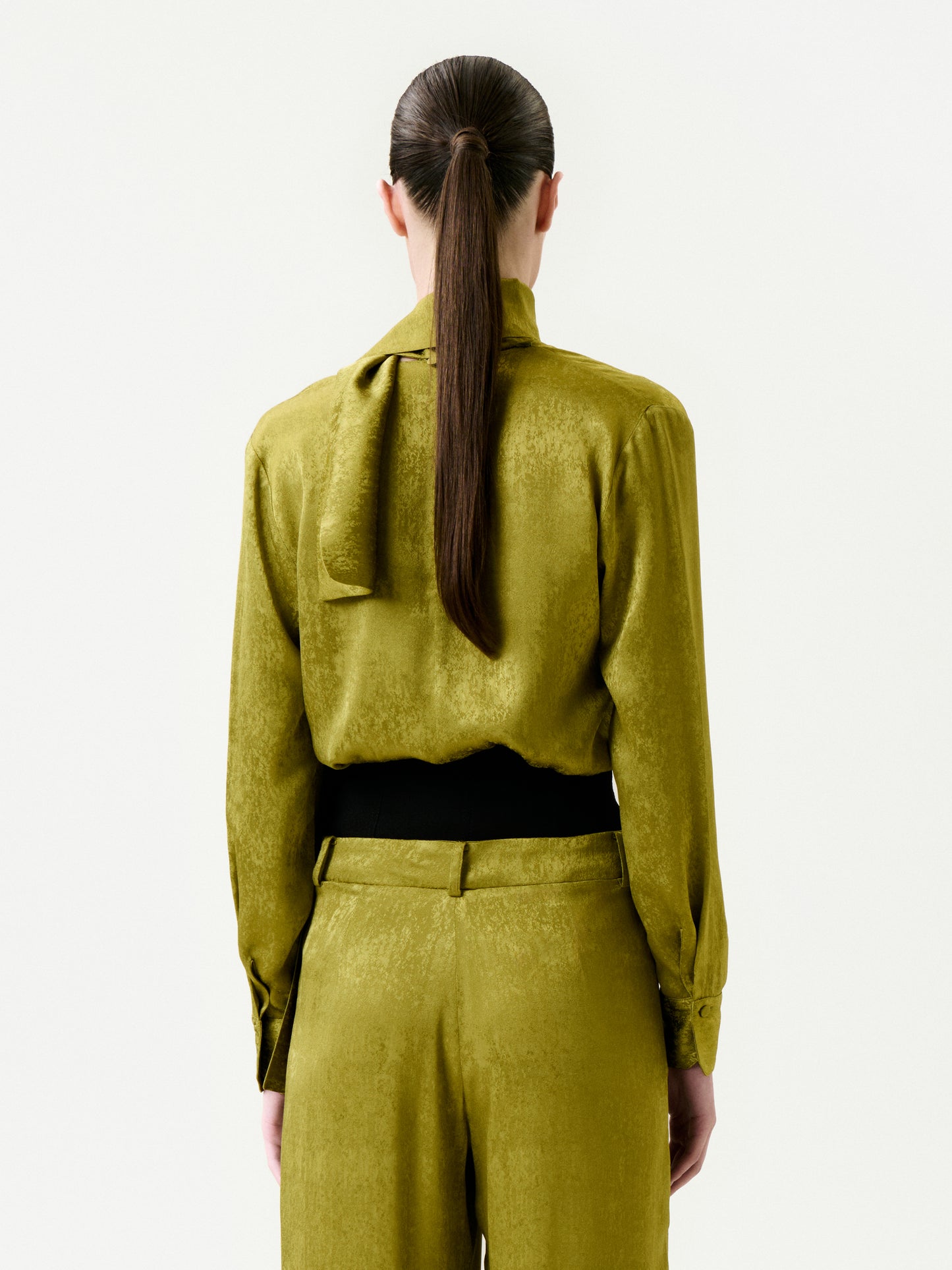 A person with long brown hair in a low ponytail is shown from behind wearing the Omara Blouse—an olive green, Italian fabric top with a neck tie—paired with matching pants, set against a plain white background.