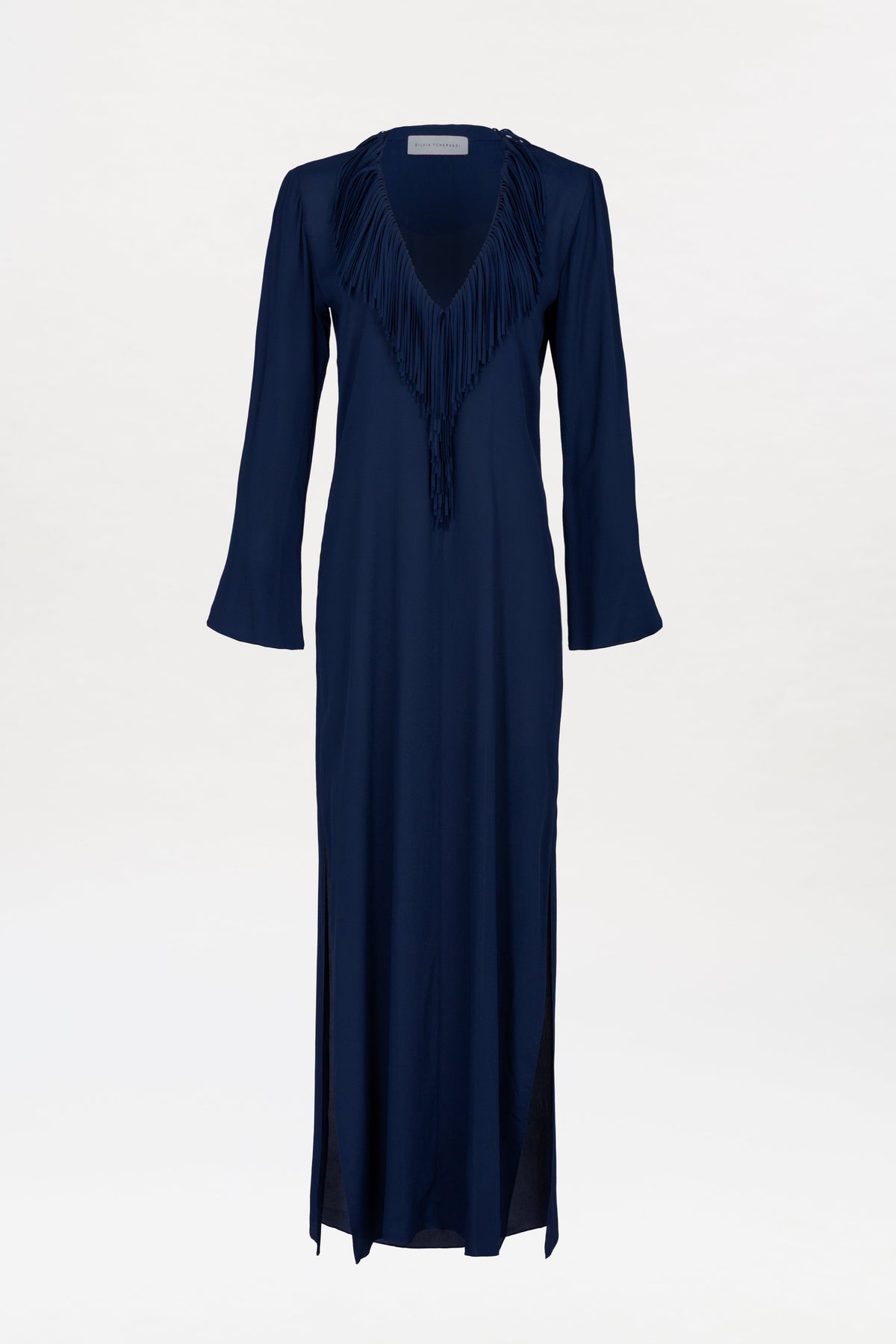 The Mercedes Dress is a long-sleeved, navy blue maxi dress featuring a deep V-neckline and fringe detailing around the collar, shown against a plain white background.