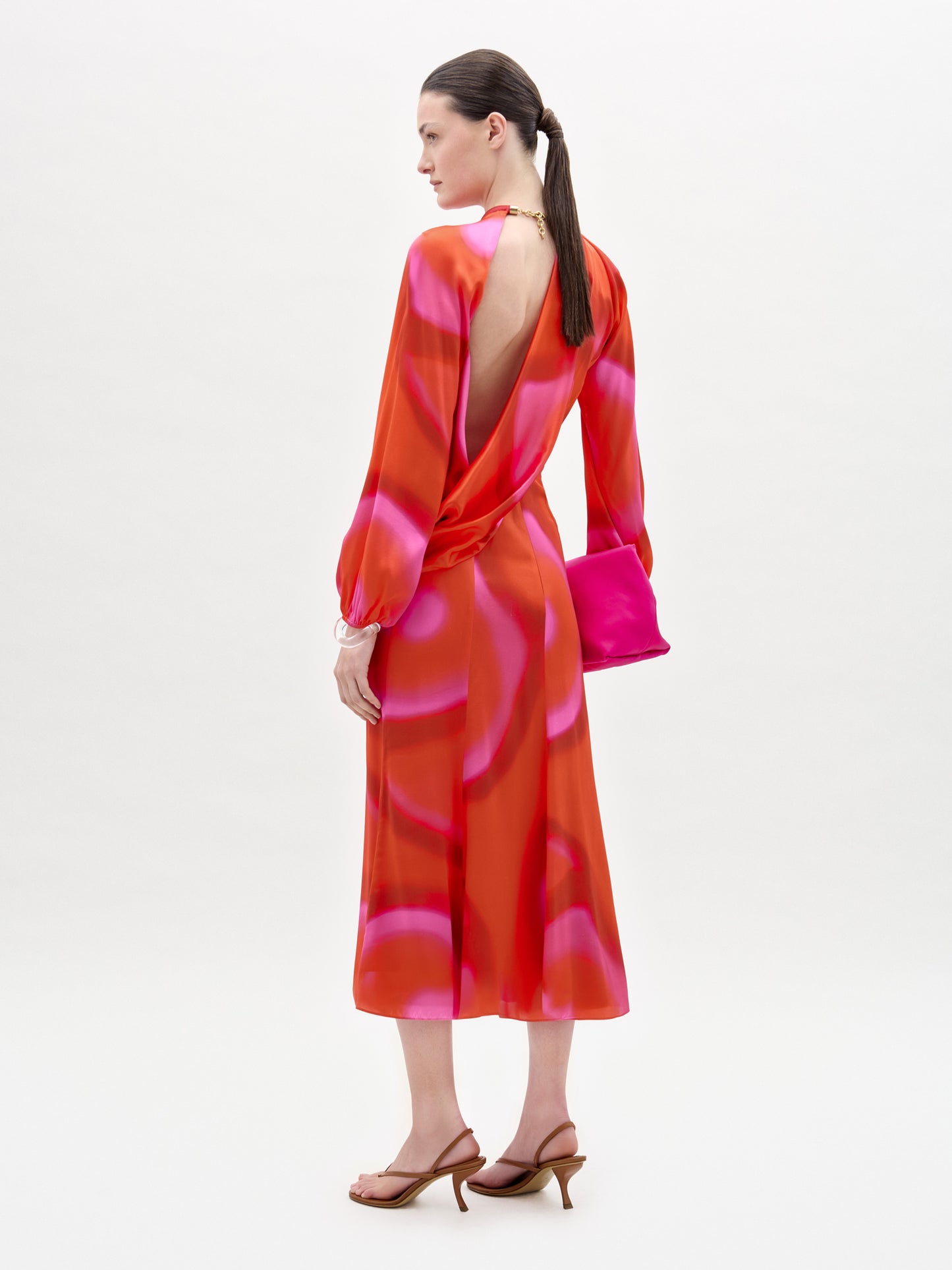 A woman faces away, wearing the Muna Dress—a red and pink patterned Italian fabric midi with open back and draped bodice—styled with brown heeled sandals and a bright pink clutch against a plain white background.