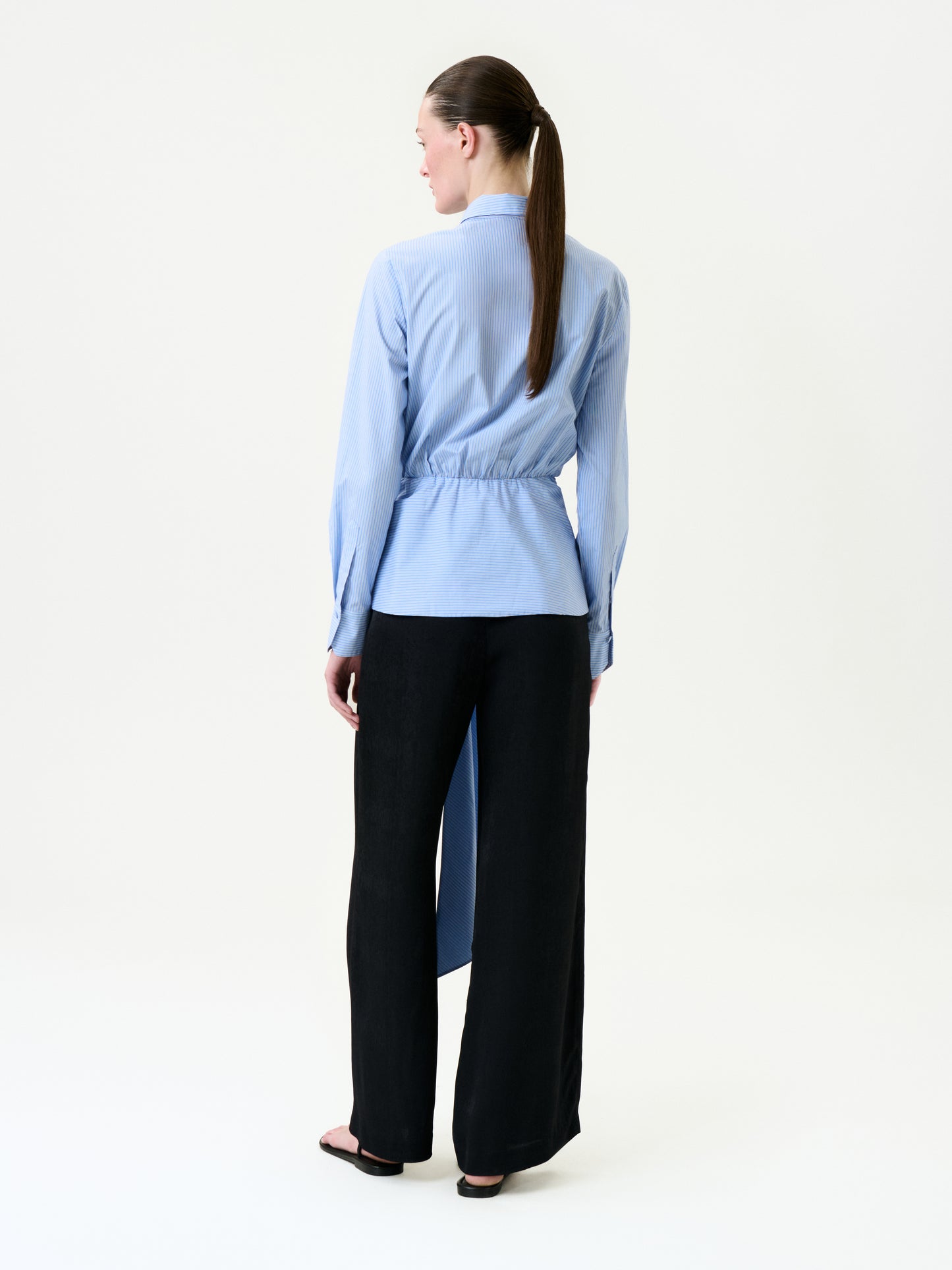 A woman with her brown hair in a low ponytail faces away, wearing loose black Ulla Pant trousers, a light blue long-sleeve 100% viscose shirt with cinched waist and asymmetrical hem, and black sandals.