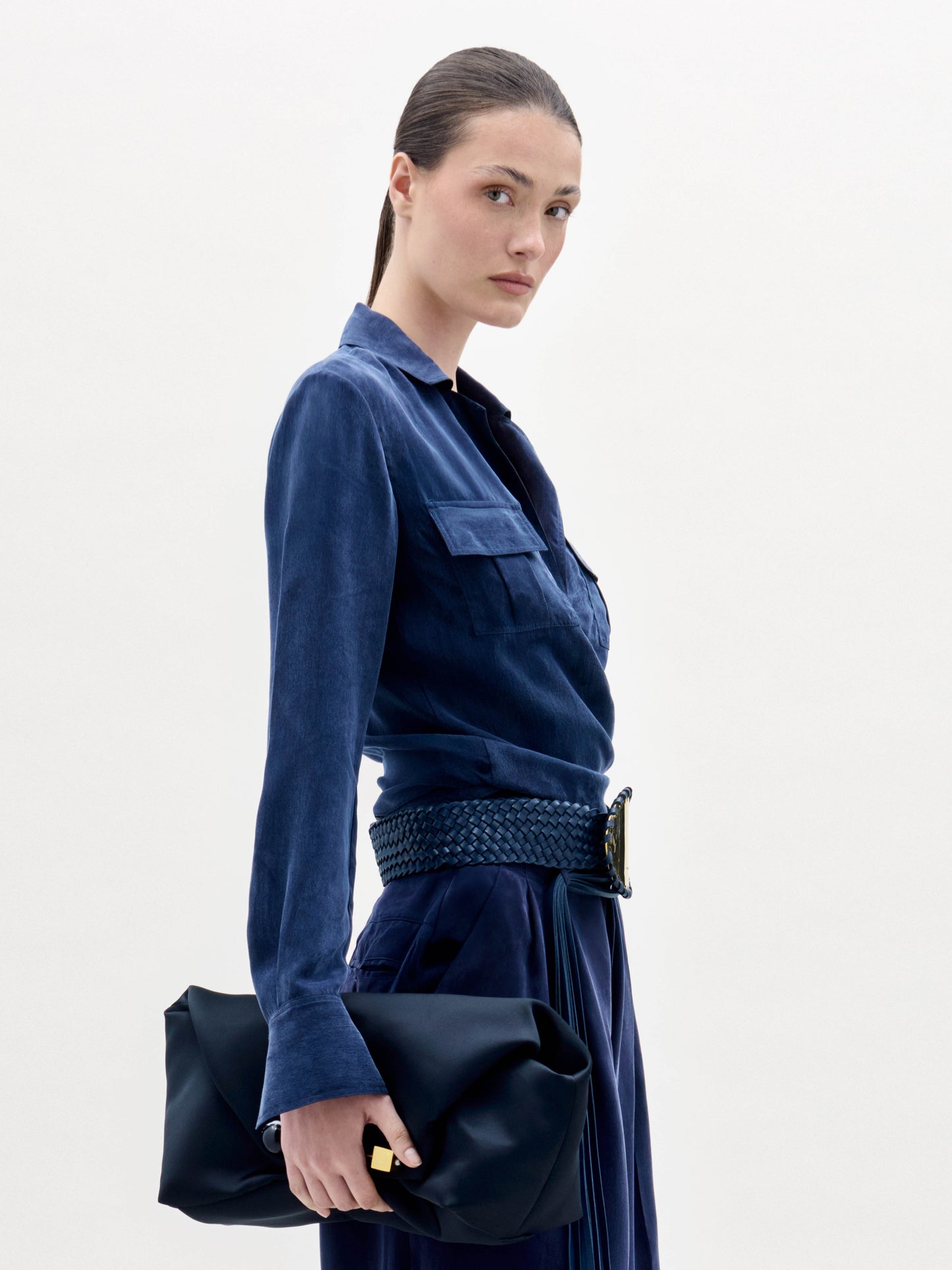 A woman wearing the Caserta Blouse in deep blue, paired with trousers, a gold-buckled belt, and a matching navy clutch, stands against a plain light background.