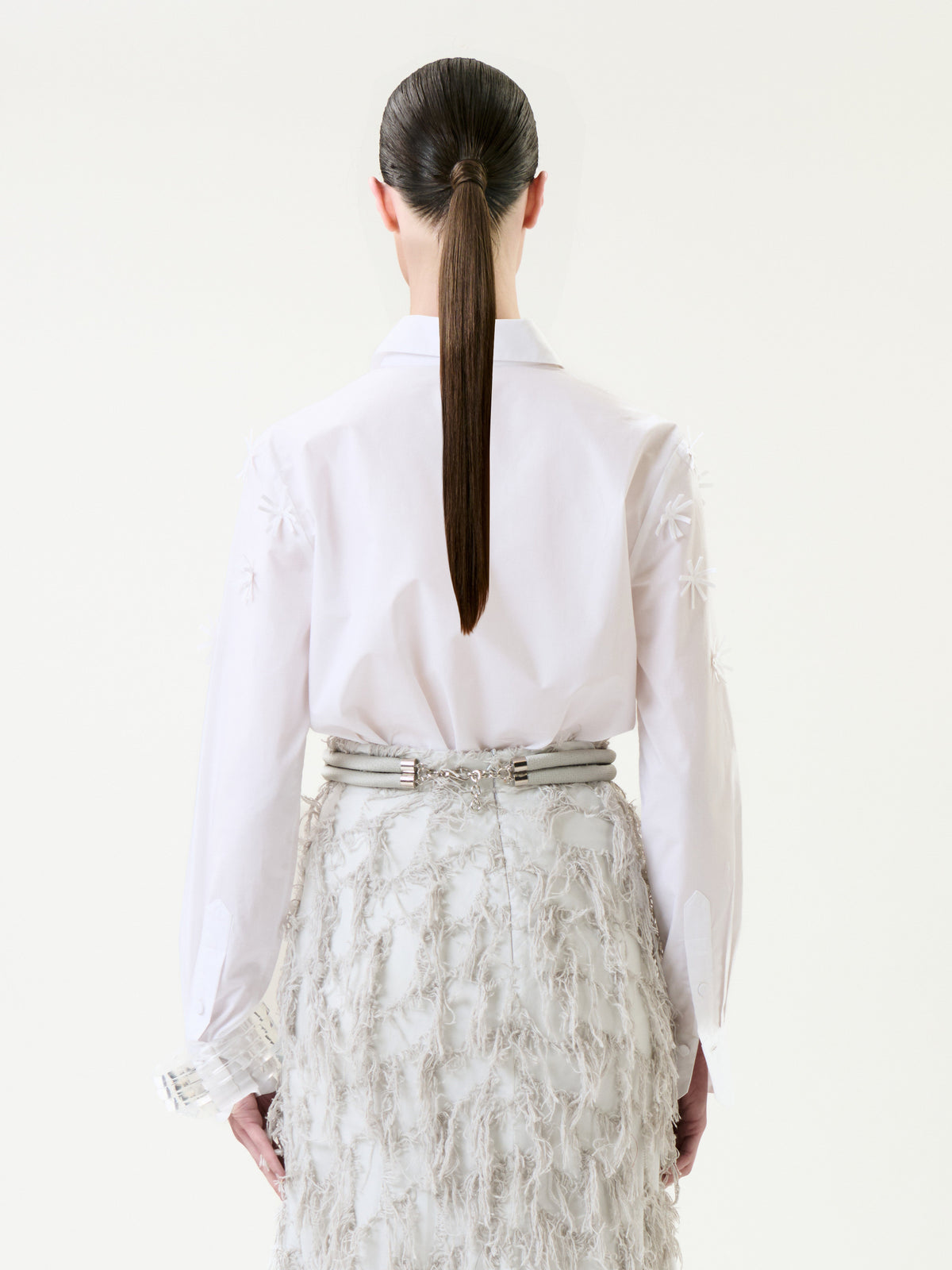 A person with long, straight brown hair in a ponytail is seen from behind against a plain background, wearing the Naroa Shirt—a white top featuring textured patterns—paired with a high-waisted, feathered white skirt.