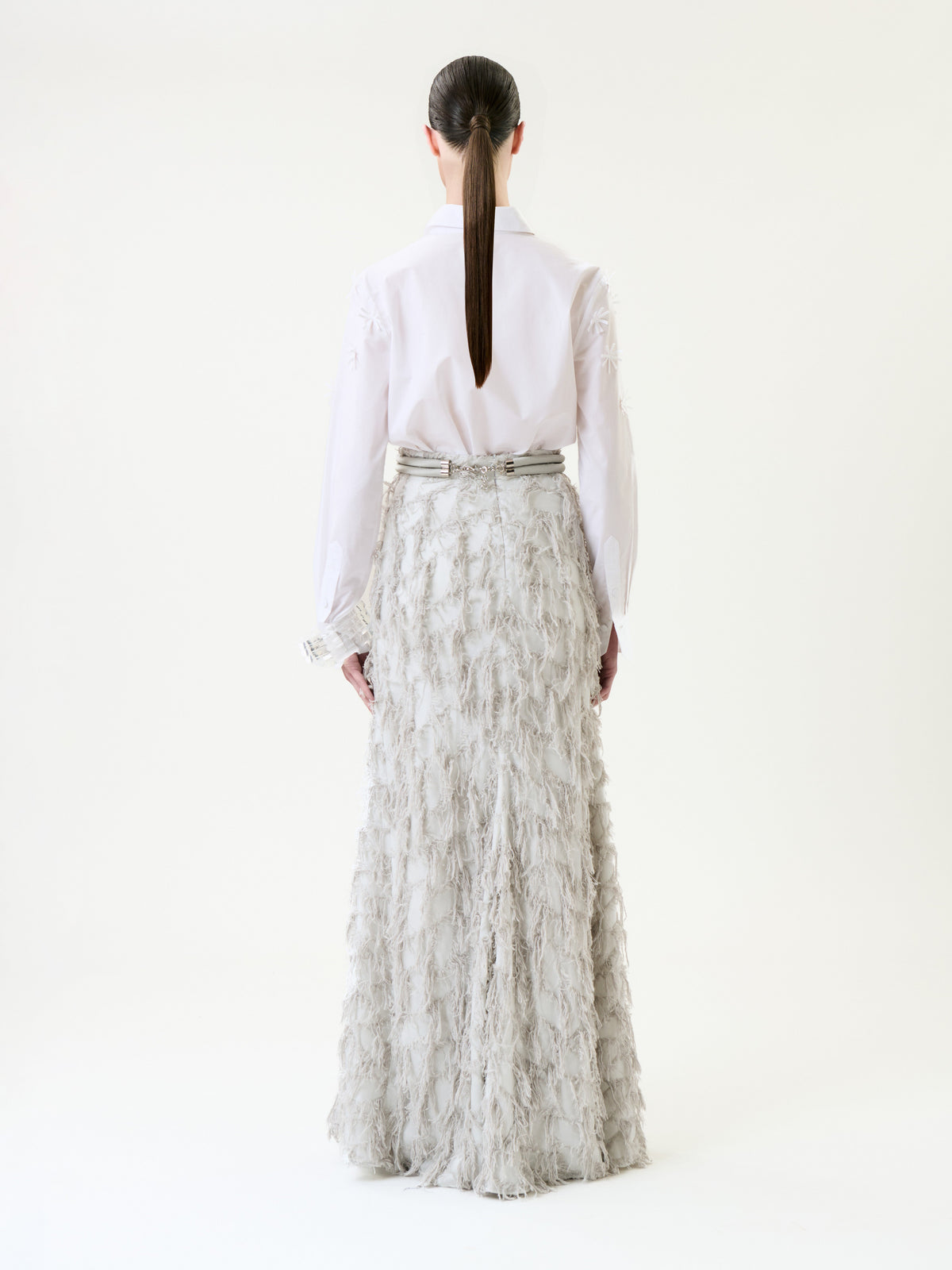 A person with long, dark hair in a low ponytail, wearing a white shirt and the Vania Skirt—a long, textured light gray skirt—stands facing away against a plain white background.