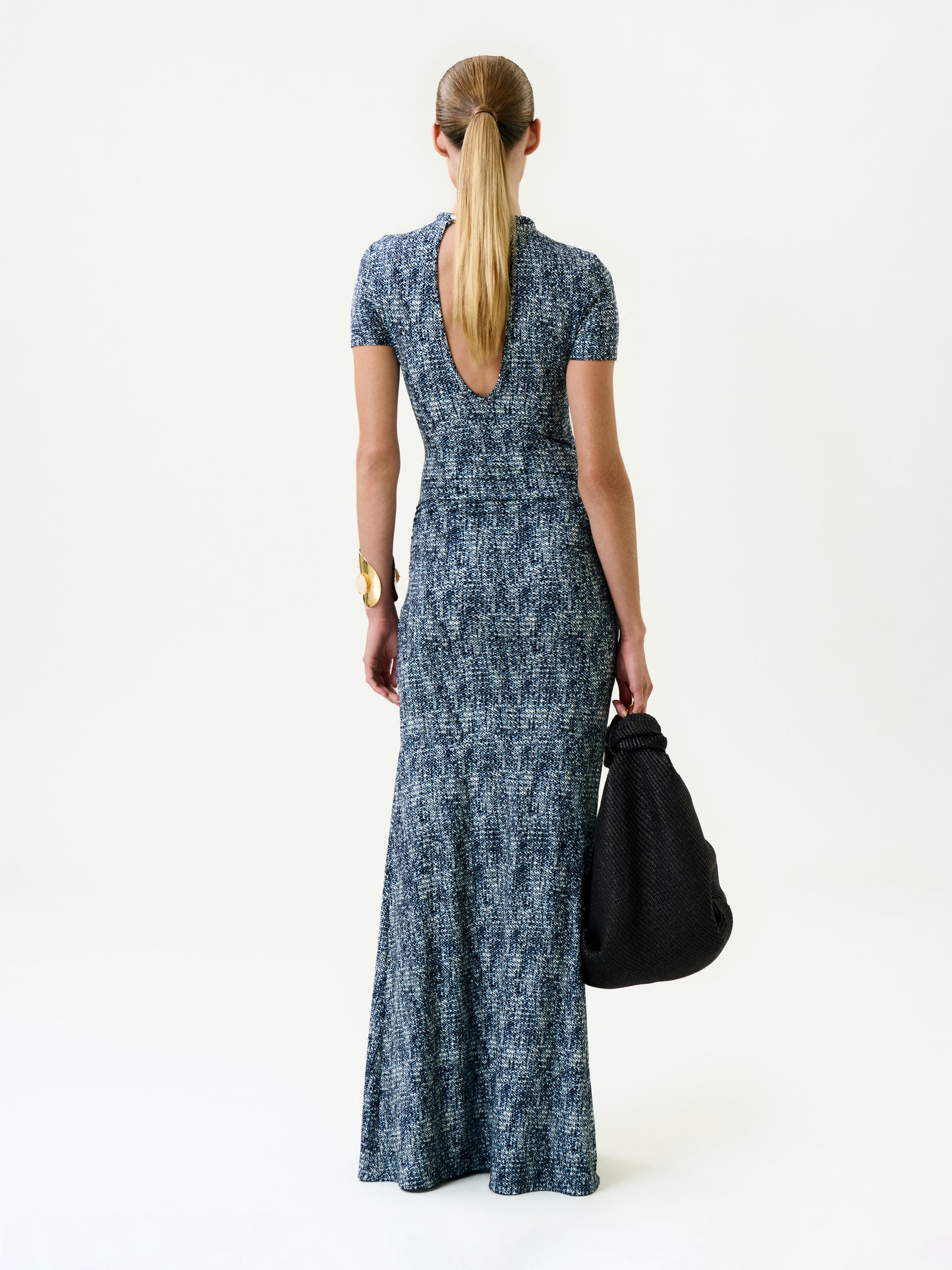 A woman with long hair in a low ponytail wears the Yara Dress, a short-sleeved blue patterned maxi from Colombia with a low scoop back. She faces away, holding a large textured black bag against a plain white background.