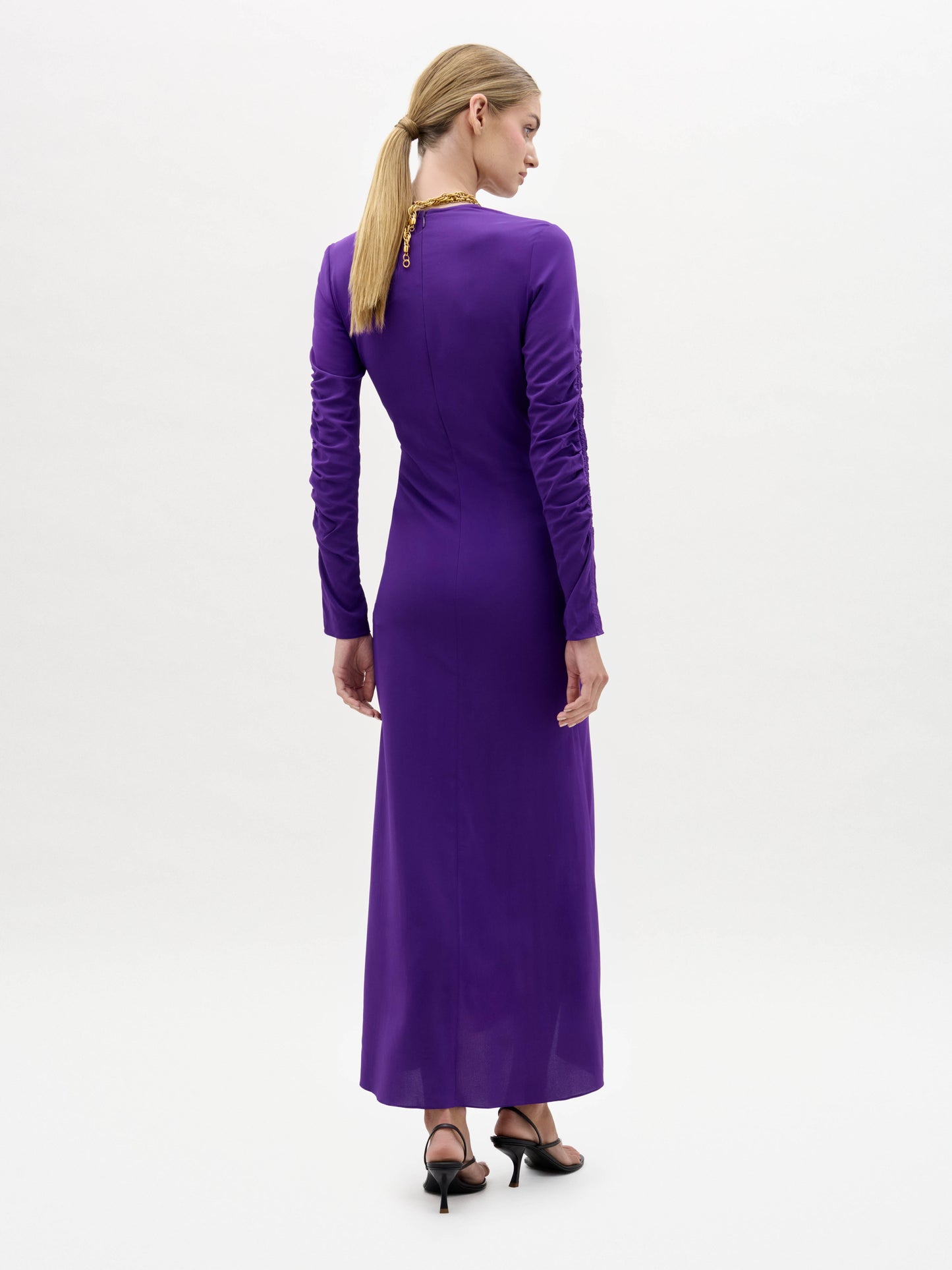 A woman stands against a plain white background, wearing the Mara Dress—a long-sleeved, asymmetrical purple dress with ruching—paired with black sandals and a long gold necklace.