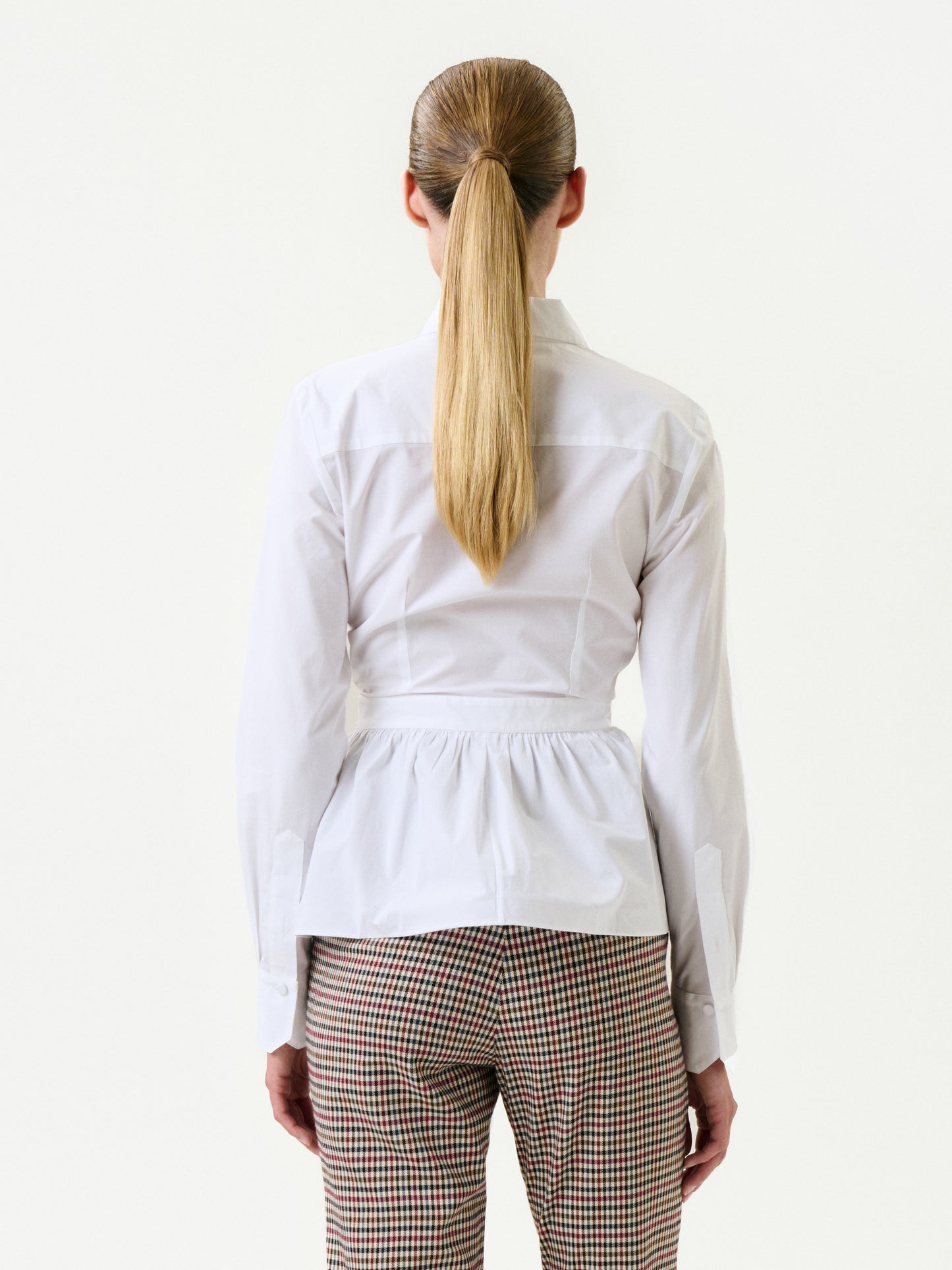 A person with long, straight, blonde hair in a ponytail is seen from behind, wearing the white Malina Shirt (100% cotton) and high-waisted brown and white checkered pants made from Portuguese fabric, set against a plain white background.