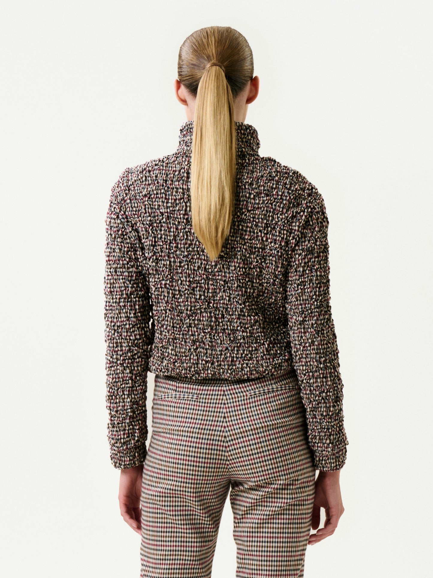 A person with long, straight blonde hair in a ponytail is shown from the back wearing the Zalta Jacket over a textured high-neck sweater and plaid pants, standing against a plain white background.