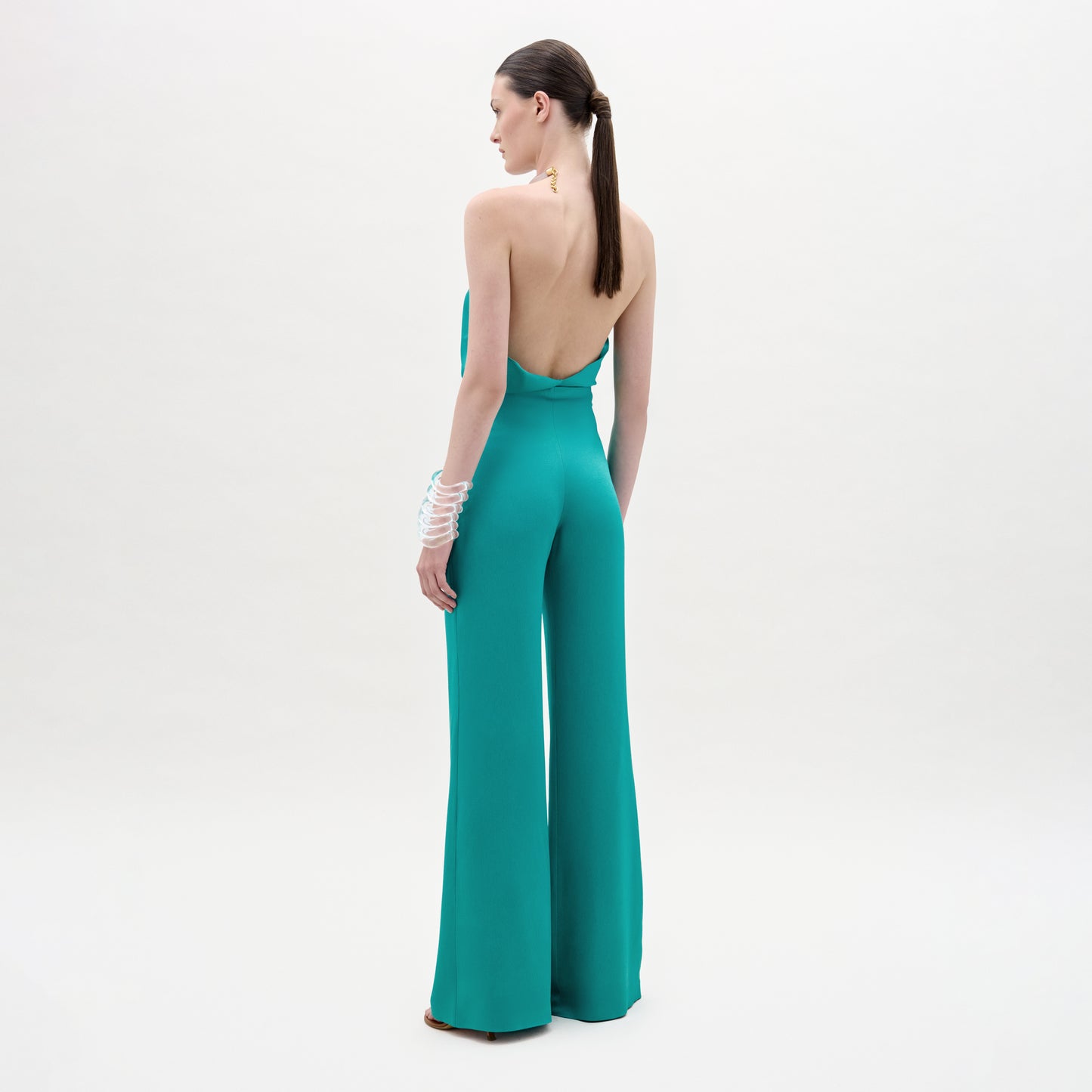 A woman faces away, wearing the teal Palermo Pant, a backless halter-neck jumpsuit with wide-leg pants made from Italian fabric. Her hair is tied back and she accessorizes with white bangles against a pale background.