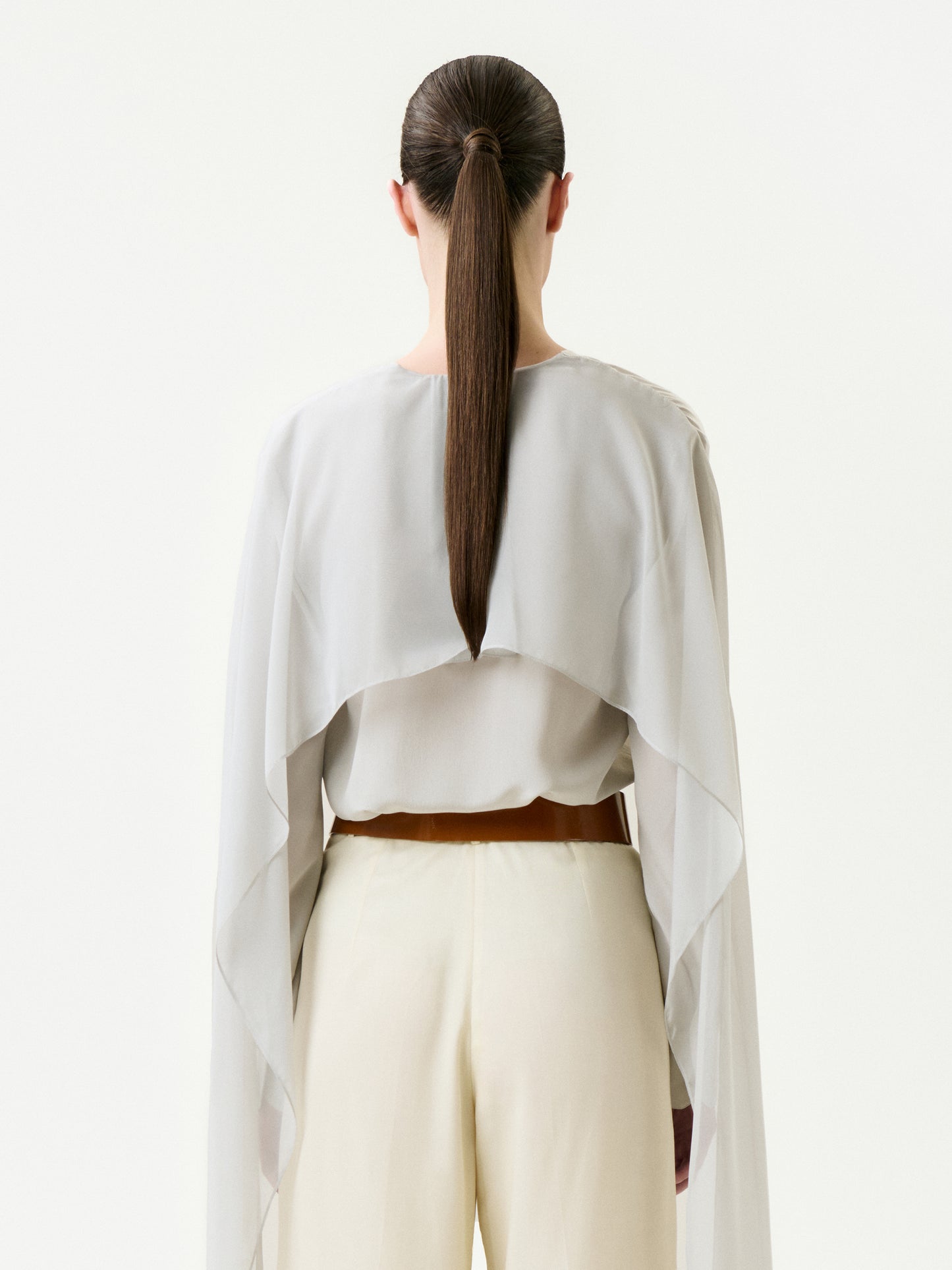 A person with long brown hair in a low ponytail is seen from the back wearing the Vanna Blouse, a light, flowy, sheer top crafted from 100% silk Italian fabric with wide sleeves, paired with cream pants against a plain white background.