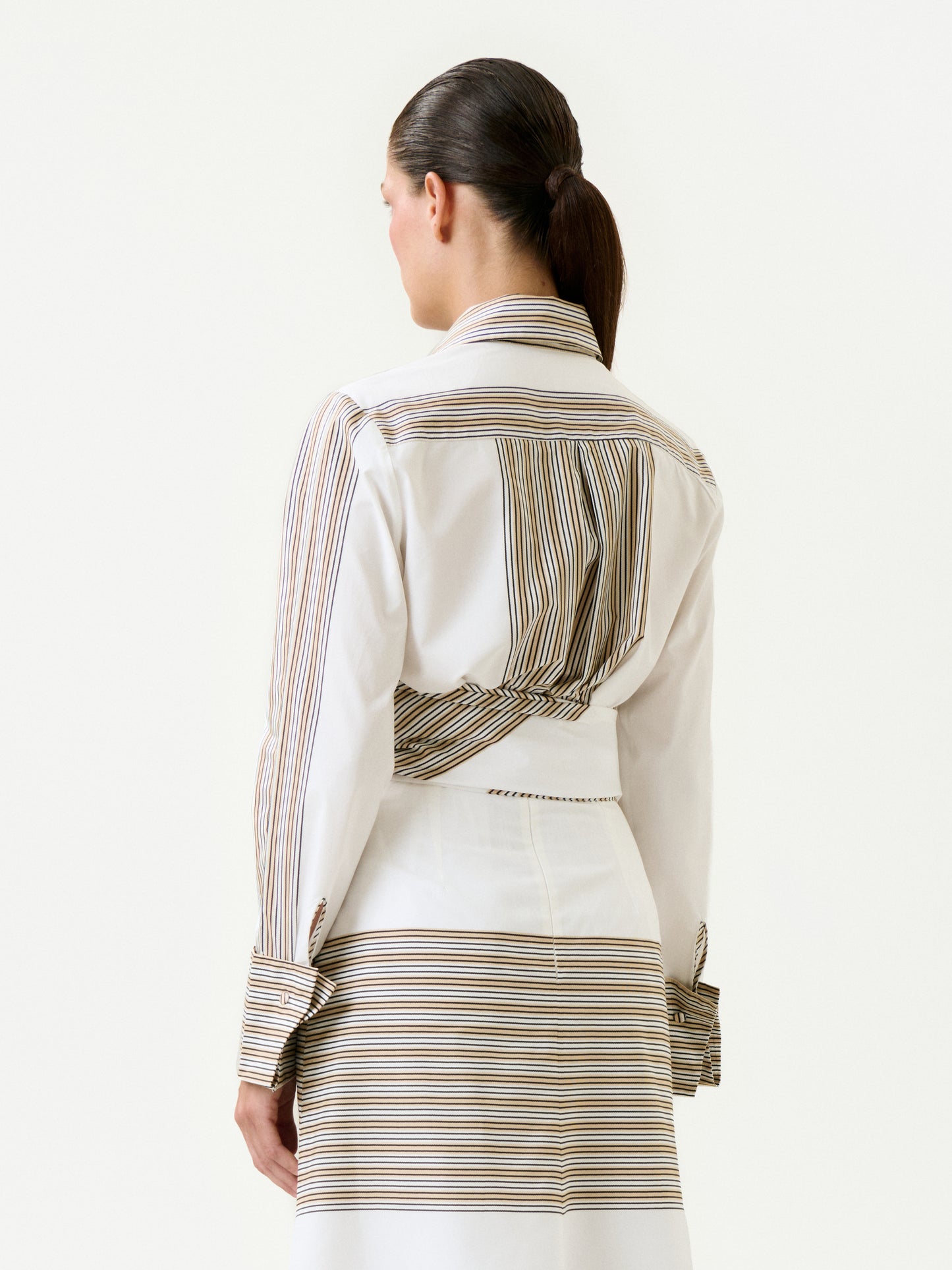 A woman with dark hair in a low ponytail is shown from the back, wearing the 100% cotton Angeles Shirt in soft Italian fabric with stripes and a matching skirt against a plain, light background.