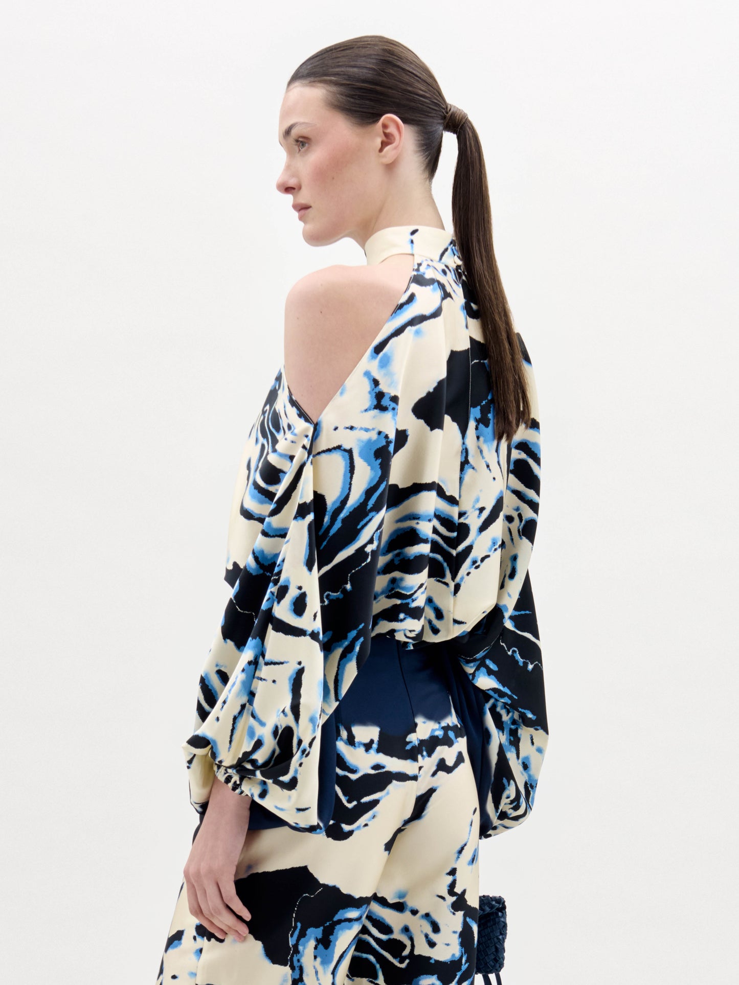 A woman stands against a plain background wearing the Safira Blouse, a high-neck, cold-shoulder top with matching wide-leg pants in a blue, black, and white marble pattern.