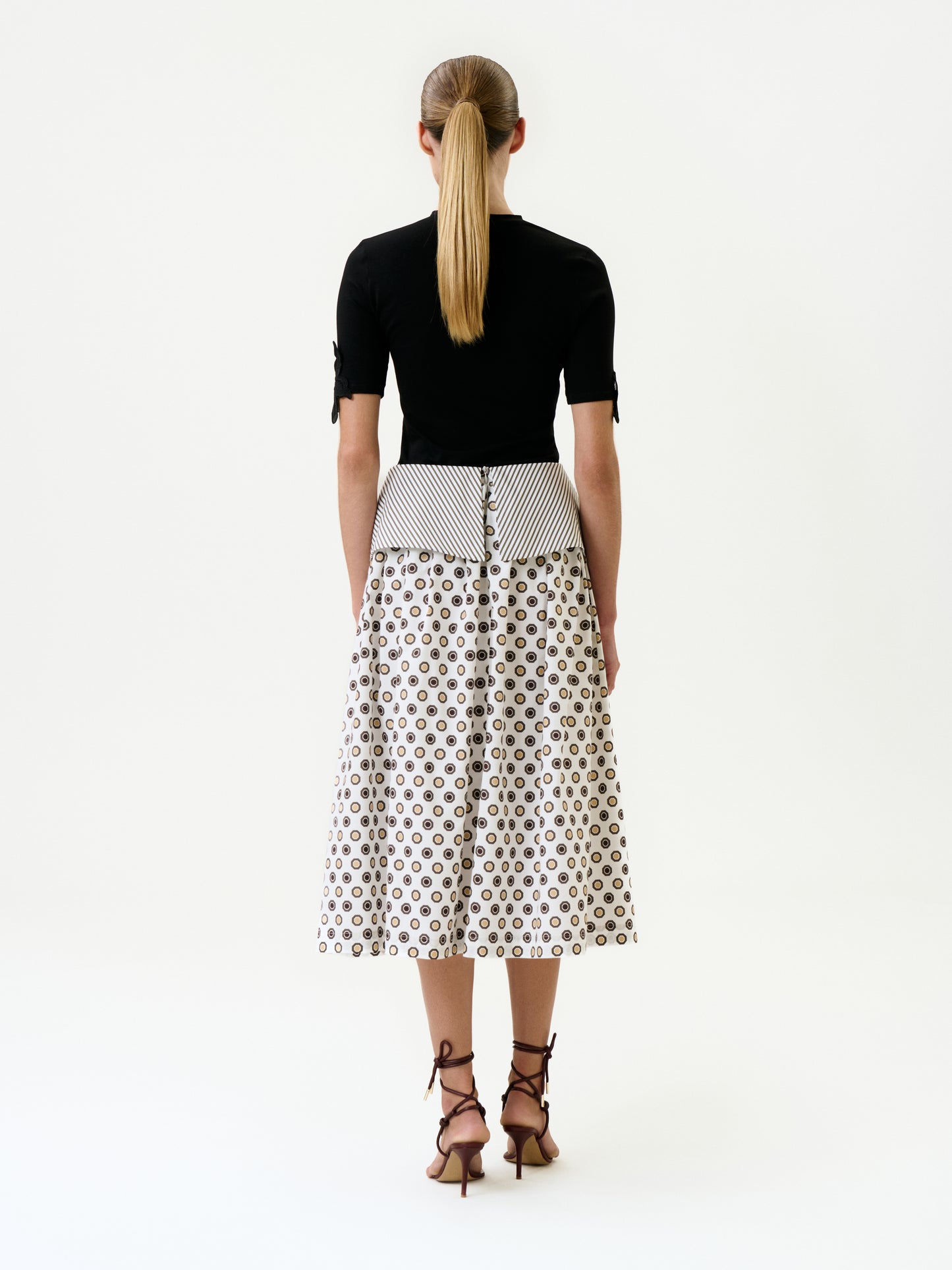 A woman with a low ponytail faces away, wearing a fitted black top and the Maxima Skirt—an elegant, high-waisted, mid-length white cotton skirt with geometric patterns. She pairs it with brown strappy heeled sandals against a plain white background.