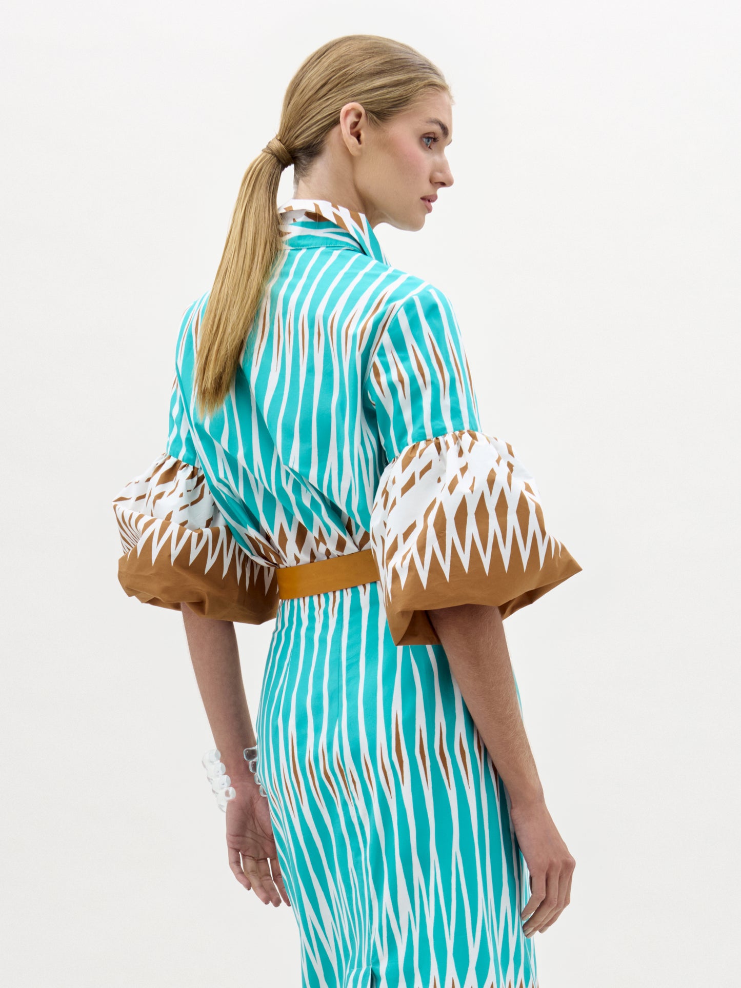 A woman with blonde hair in a ponytail models the Helina Blouse—a turquoise and brown patterned top with wide bell sleeves—standing against a plain light background, facing away with her head turned to the side.