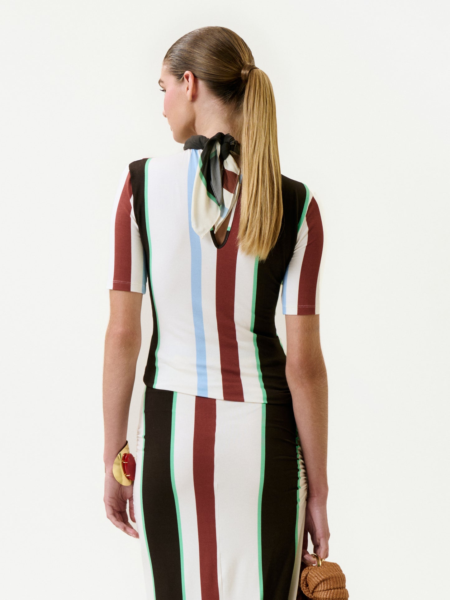 A woman with a sleek ponytail is seen from behind, wearing the Rowena Top with vertical brown, white, blue, and green stripes. She holds a small textured bag and wears a black and white scarf around her neck.