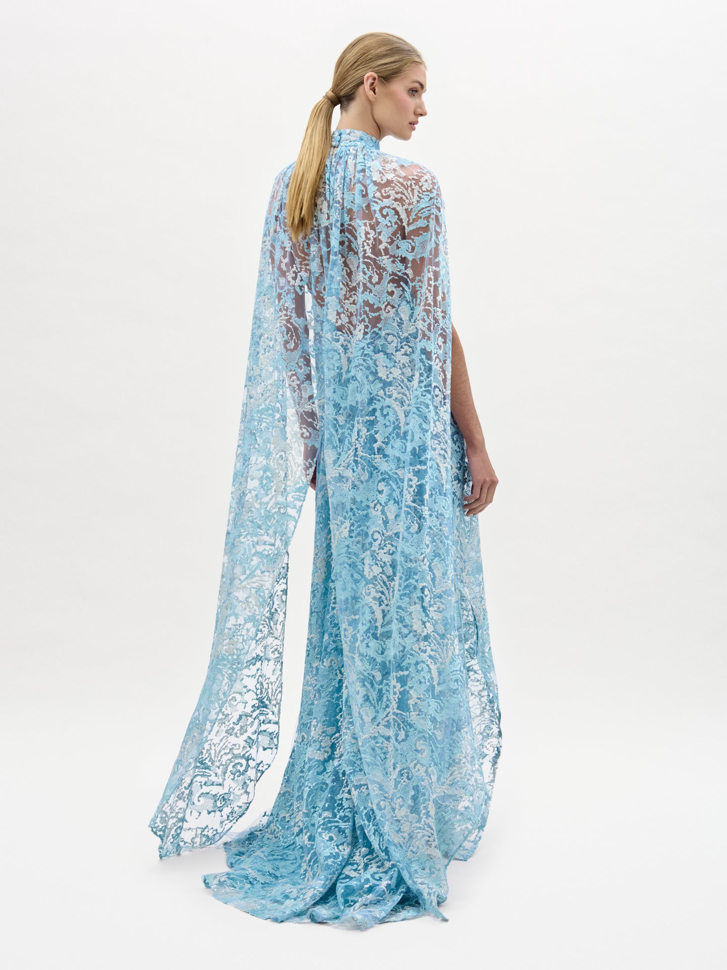 A woman stands facing away, wearing the Carol Dress—a long, flowing blue lace gown with cape-style shoulders. Her blonde hair is tied back in a low ponytail against a plain white background.