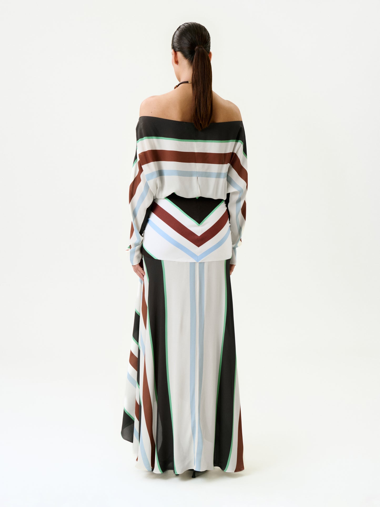 A woman with long dark hair in a low ponytail stands facing away, wearing the Sara Dress—a long, off-the-shoulder maxi dress with wide white, brown, black, blue, and green stripes—made in Colombia.