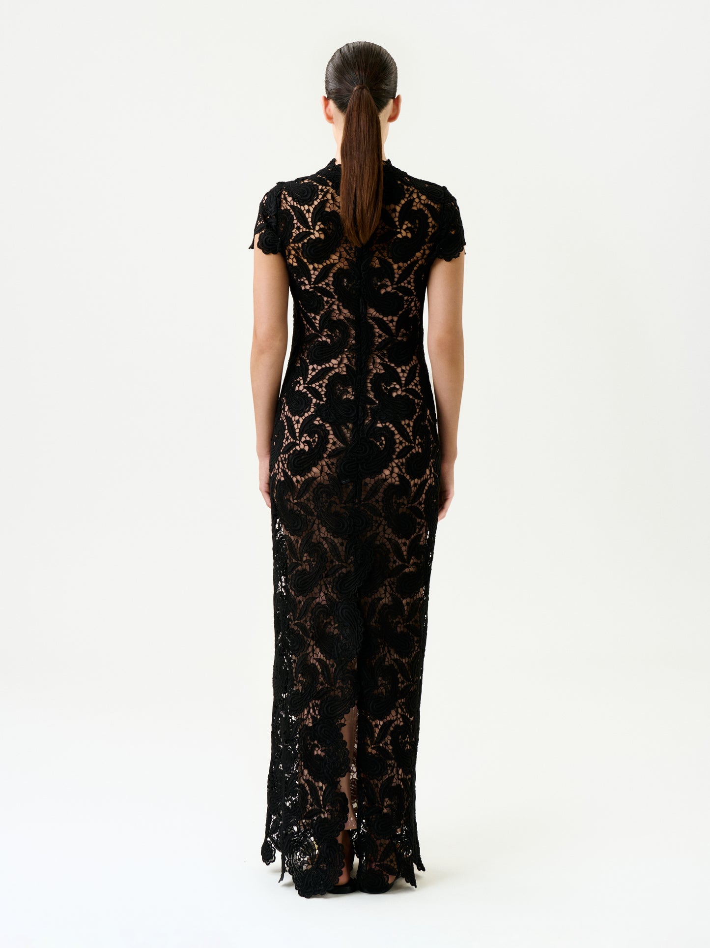 A woman with straight brown hair in a low ponytail stands facing away, wearing the fitted, floor-length Roxana Dress—a black lace maxi with short sleeves and intricate patterns—crafted from luxurious Italian fabric against a plain light background.