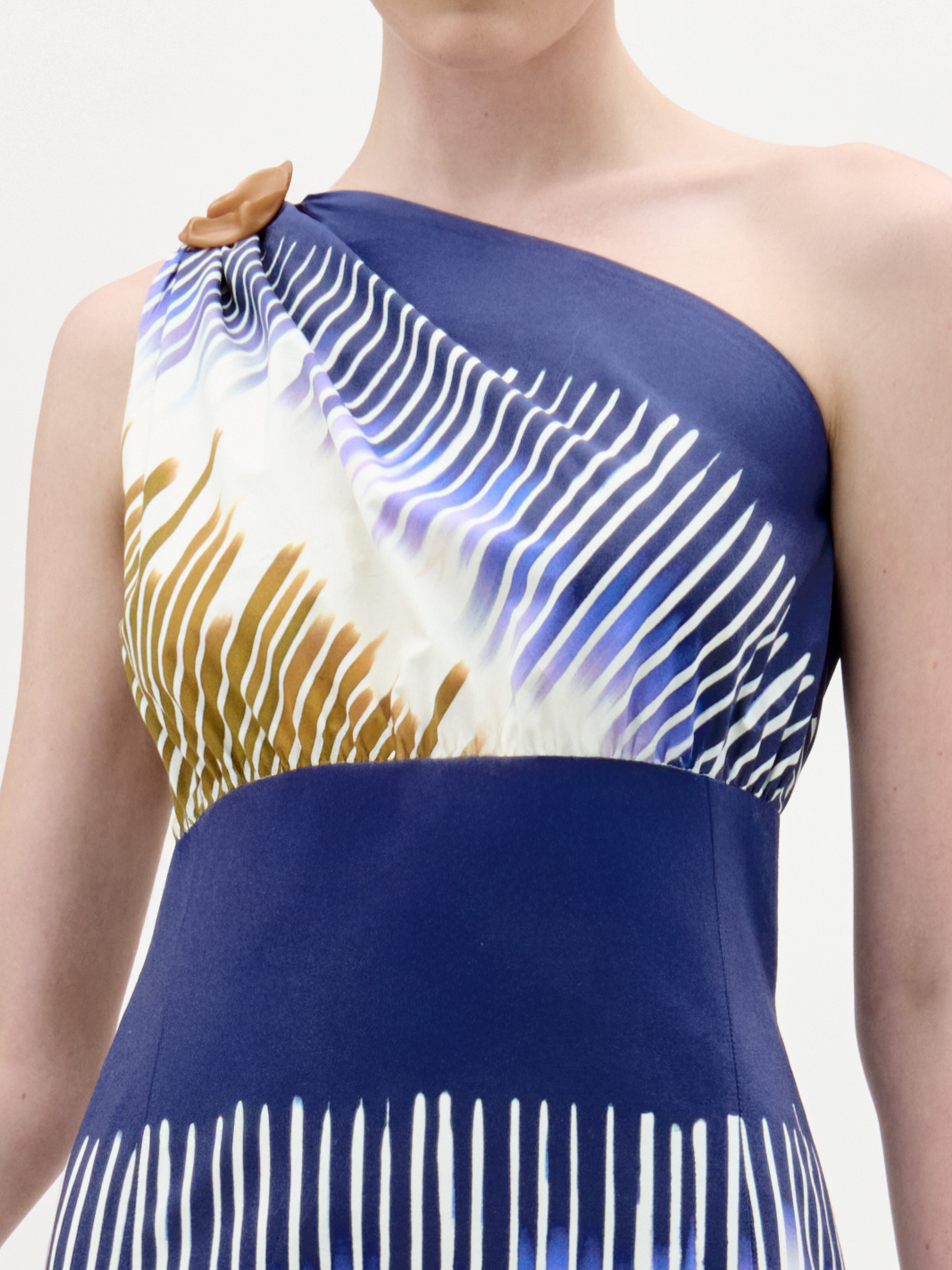 A person wearing the Catalina Dress, a fitted, smooth Italian-fabric midi with a blue base, white, gold, and purple diagonal stripes, a one-shoulder neckline, and a brown accent at the shoulder. The person's head is partially out of frame.