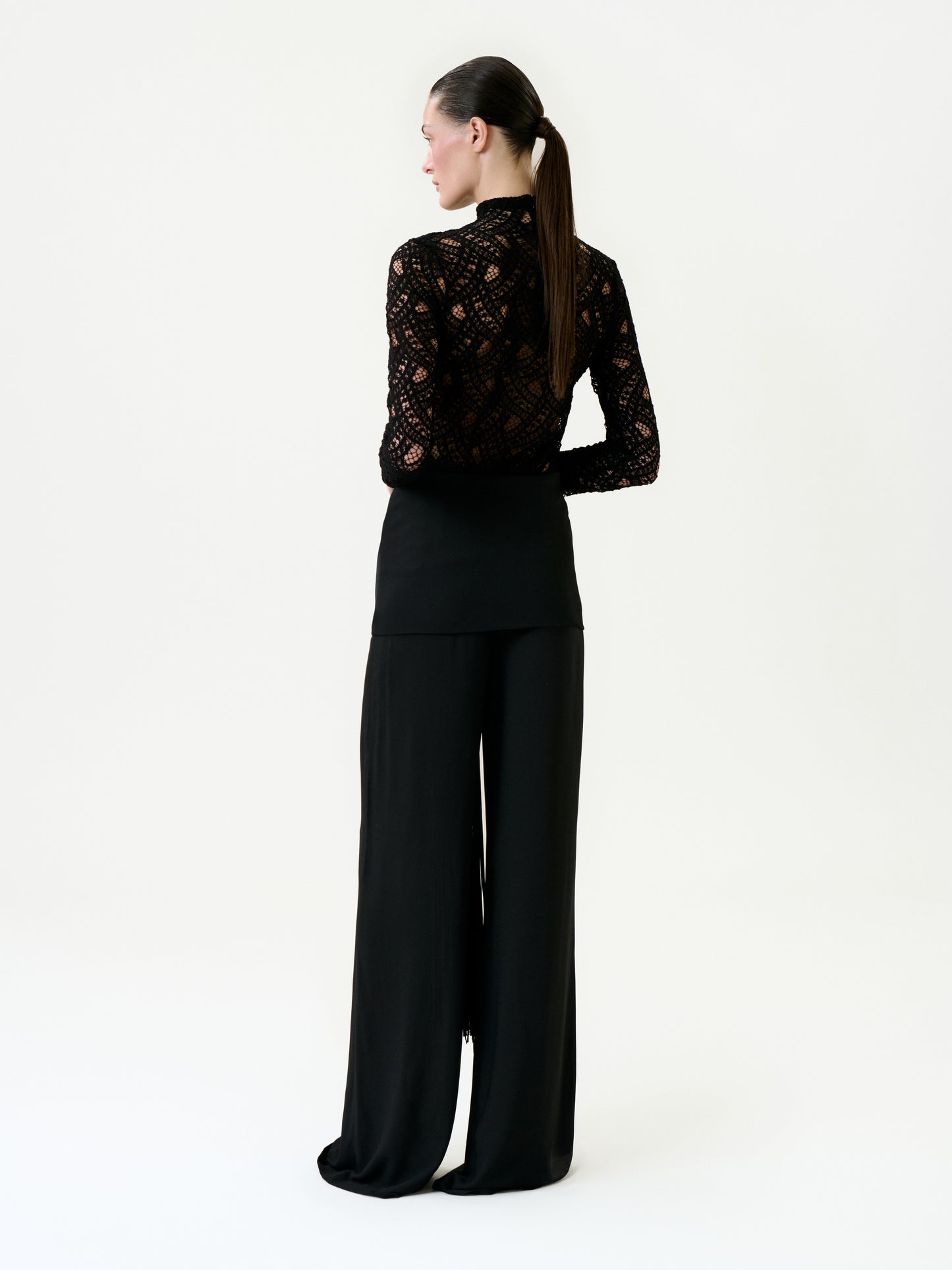 A woman faces away, styled in a black lace long-sleeve top and Kiele Pant—high-waisted, wide-leg, black pants made from premium Italian fabric. Her dark hair is tied in a low ponytail against a plain white background.