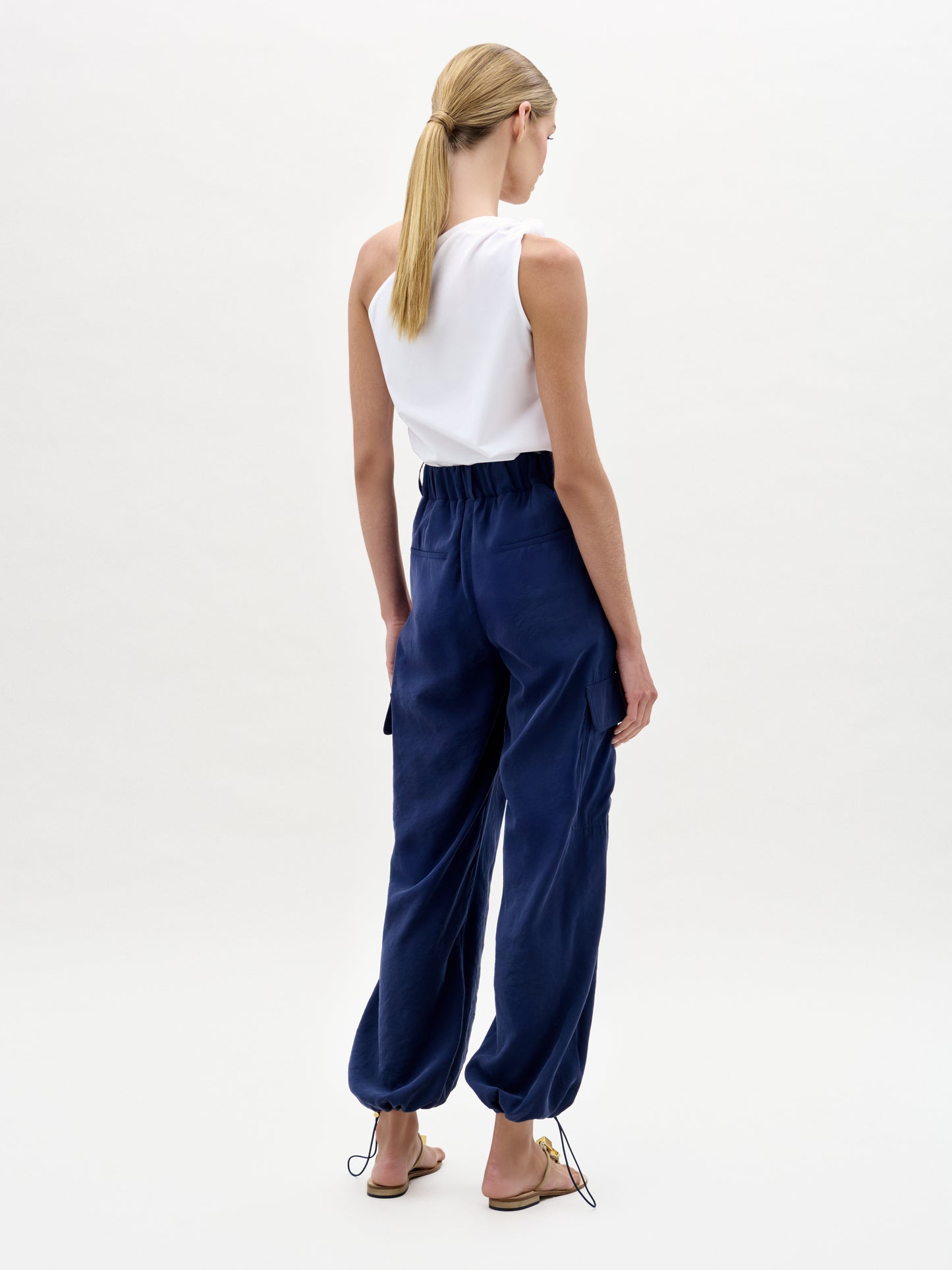 A woman with blonde hair in a low ponytail wears a sleeveless white top and the Darnell Pant—loose, high-waist navy blue cargo pants with a drawstring hem, made from fine Italian fabric. She stands facing away against a plain light background in flat sandals.