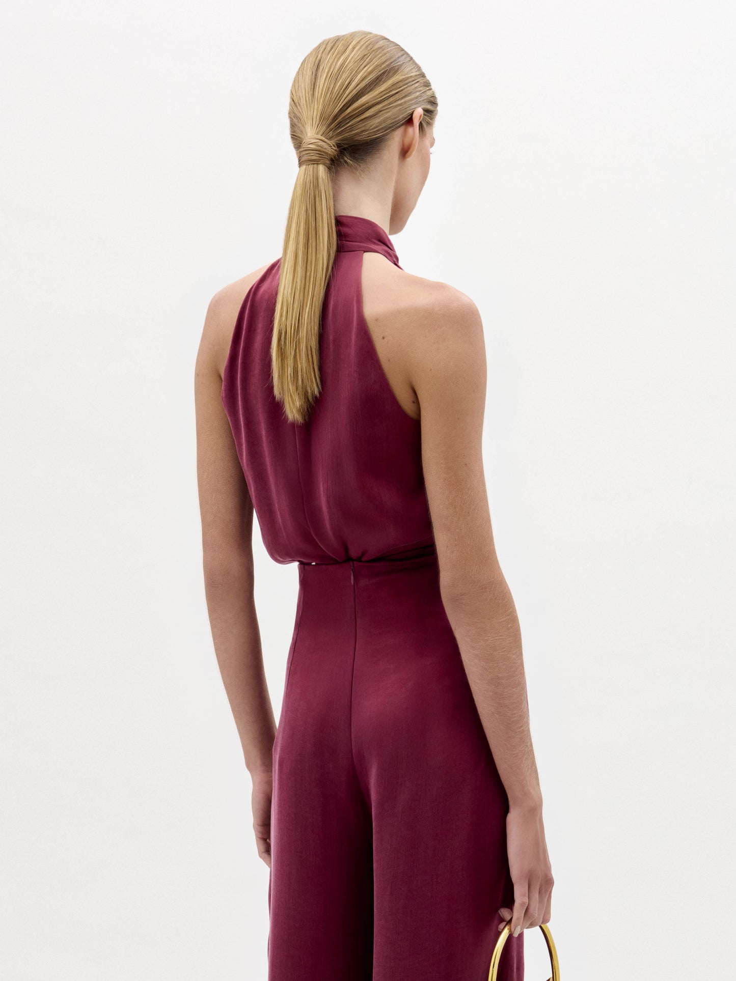 A woman stands against a plain background wearing the Shela Blouse, a sleeveless, high-neck magenta top with a tie detail at the waist, paired with a small handbag.