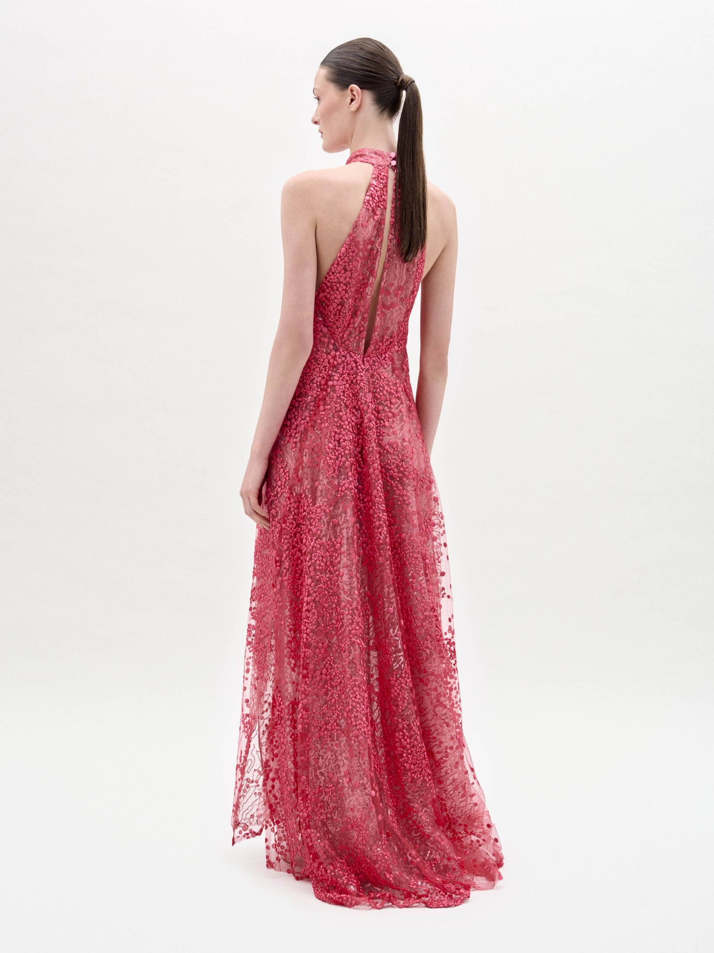 A woman stands in a studio wearing the Tatiana Dress, a long, sleeveless pink lace gown with a high neck and sheer overlay, paired with flat sandals.