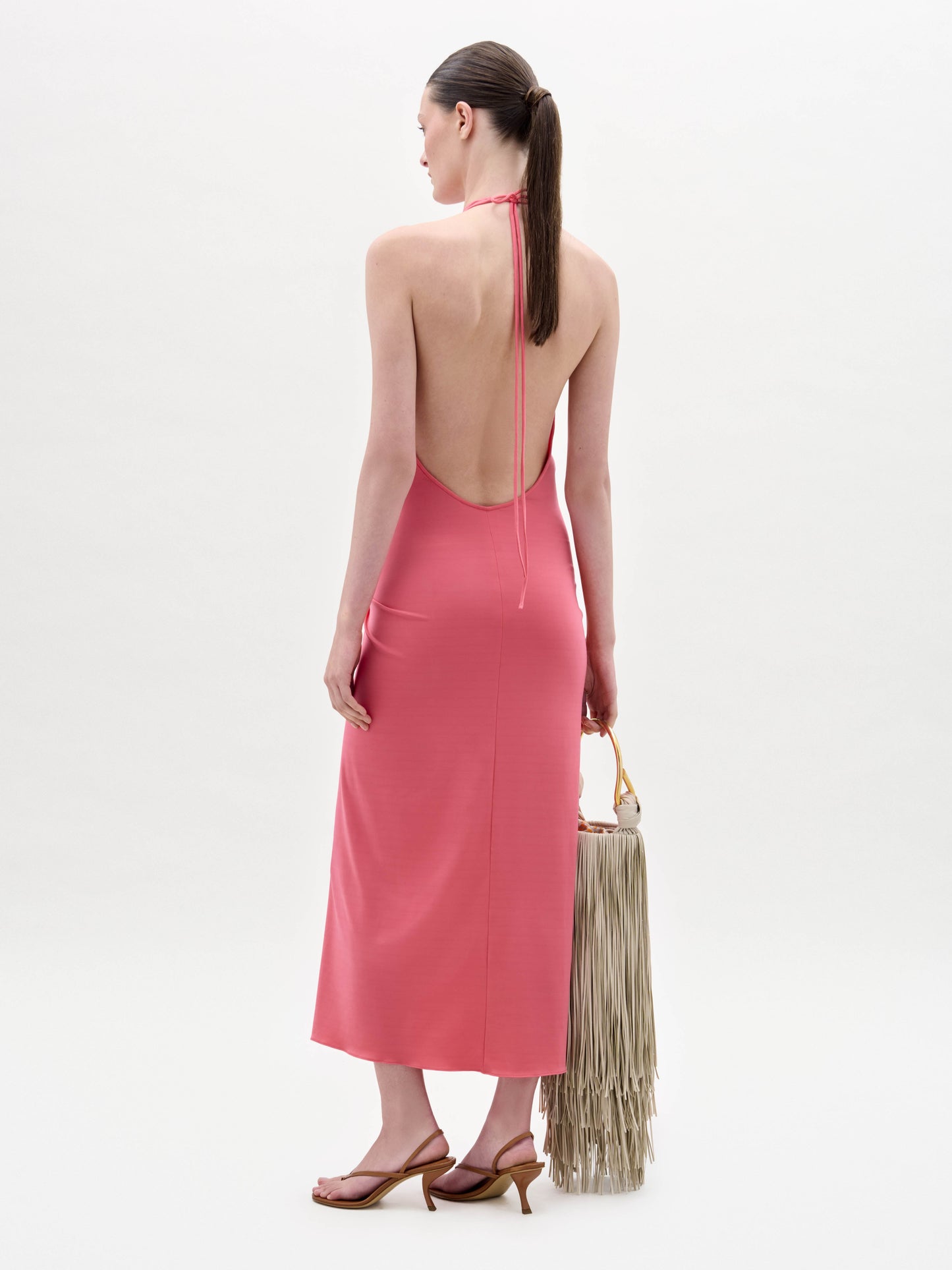 A woman stands against a plain background wearing the sleeveless pink Veracruz Dress with a gathered waist, holding a large beige fringed bag and wearing brown sandals.