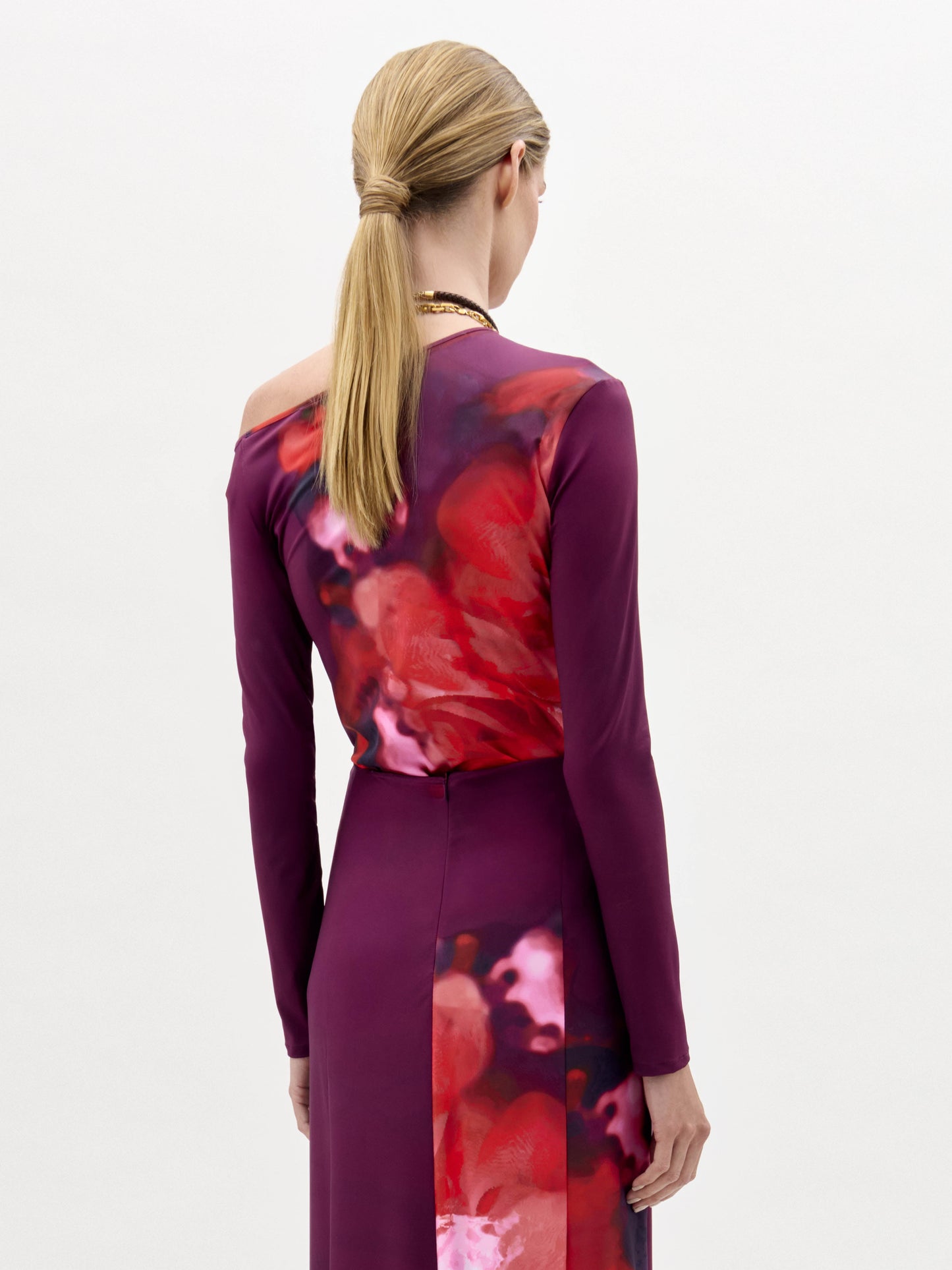 A woman wears the Sabra Blouse, a long-sleeve purple and red patterned top with an asymmetric neckline and a long gold necklace, standing against a plain white background.