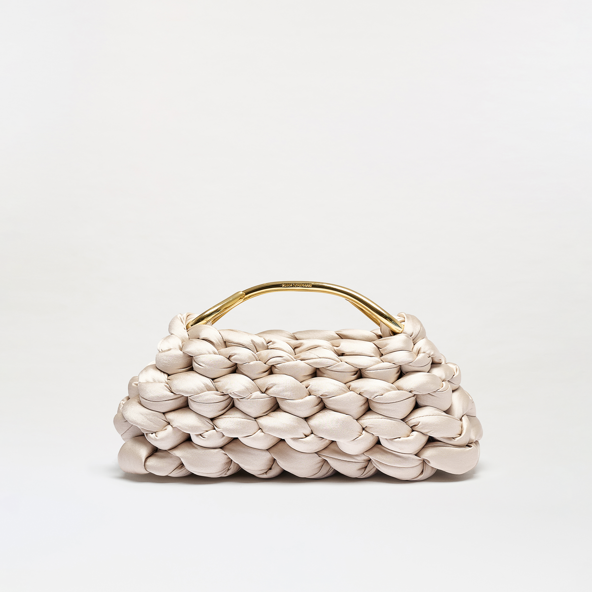A structured woven leather handbag in a light champagne beige color, featuring a curved gold-tone metal top handle, set against a clean white studio background.