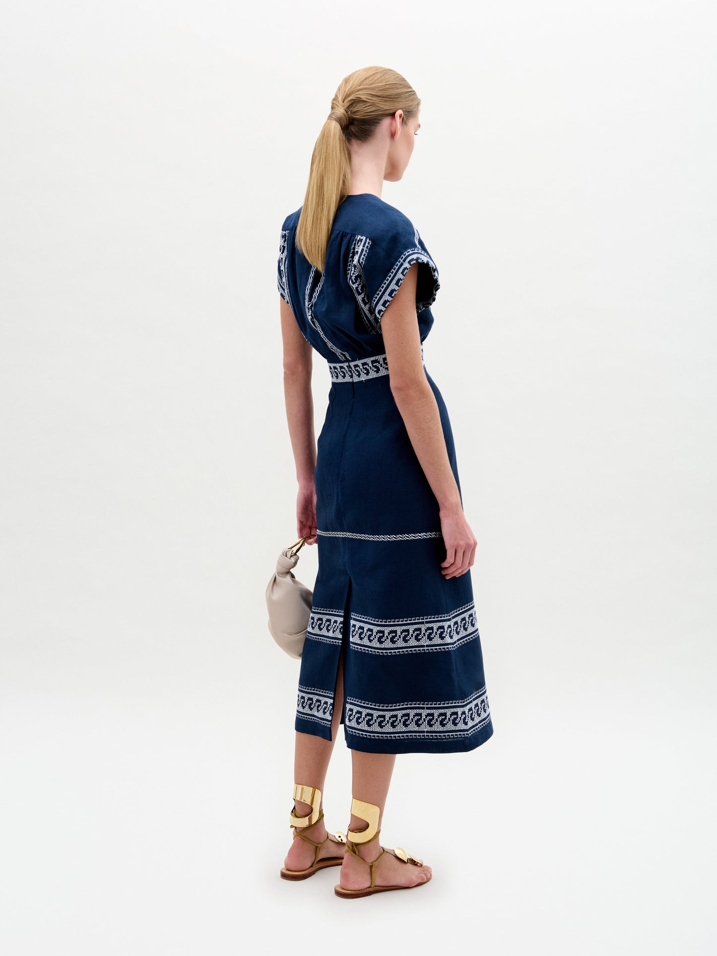 A woman stands against a plain background wearing the Ady Dress, a navy blue embroidered linen midi with white patterns, gold sandals, and holding a light-colored handbag.